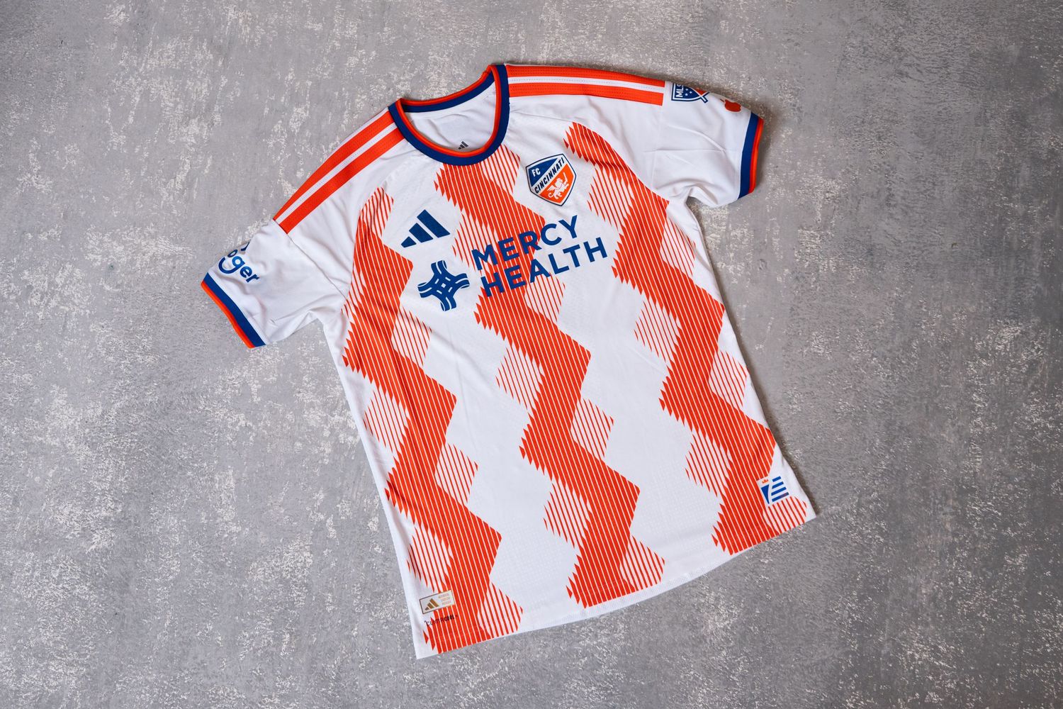 Flat-lay view of a white and orange FC Cincinnati jersey displayed on a textured gray surface.