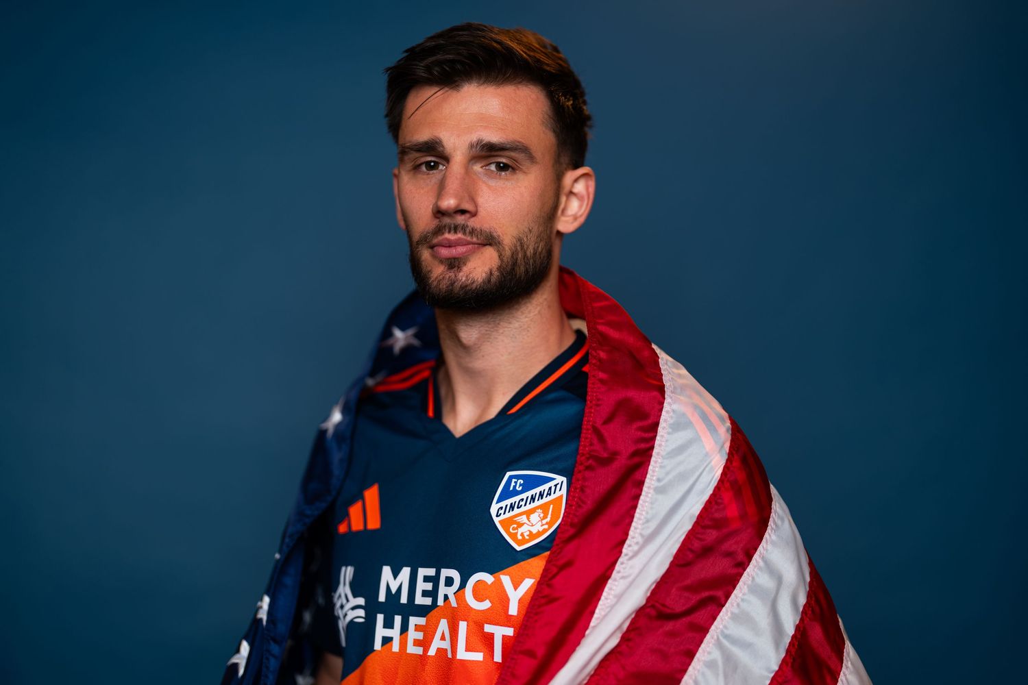 Studio portrait of a person wearing a blue and orange FC Cincinnati jersey draped in an American flag against a blue background.