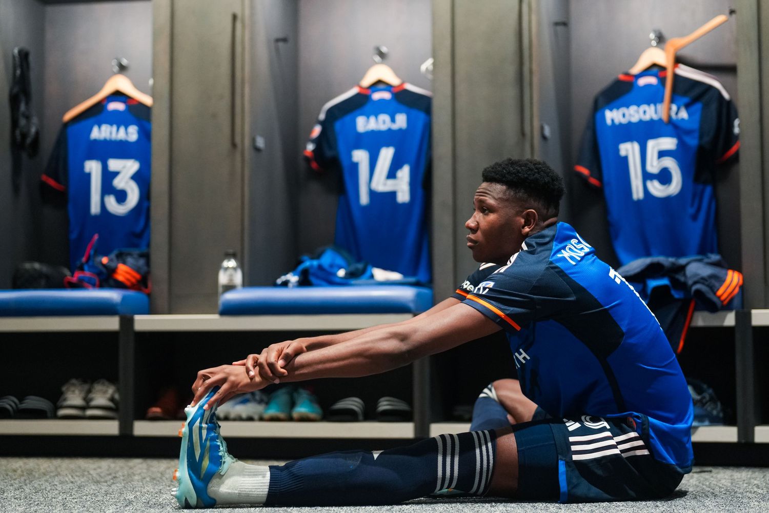 Soccer player stretches in a locker room with team jerseys hanging in the background.