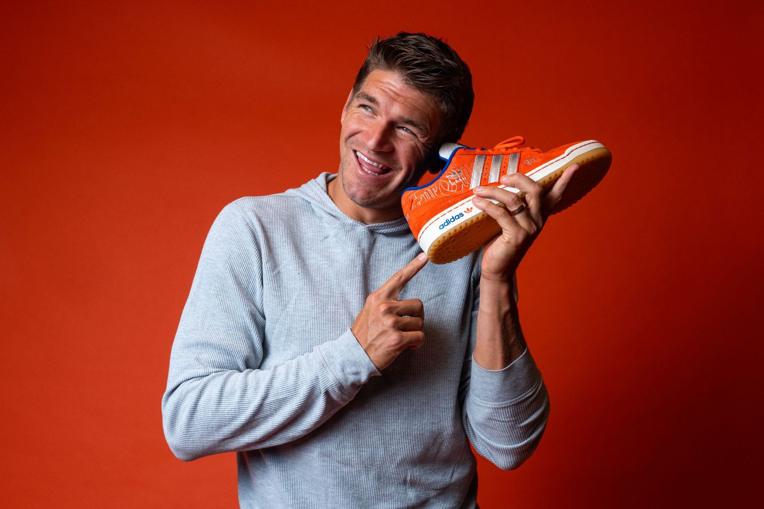 Person holding an orange Adidas sneaker to the side of their head while pointing toward the shoe against an orange studio background.