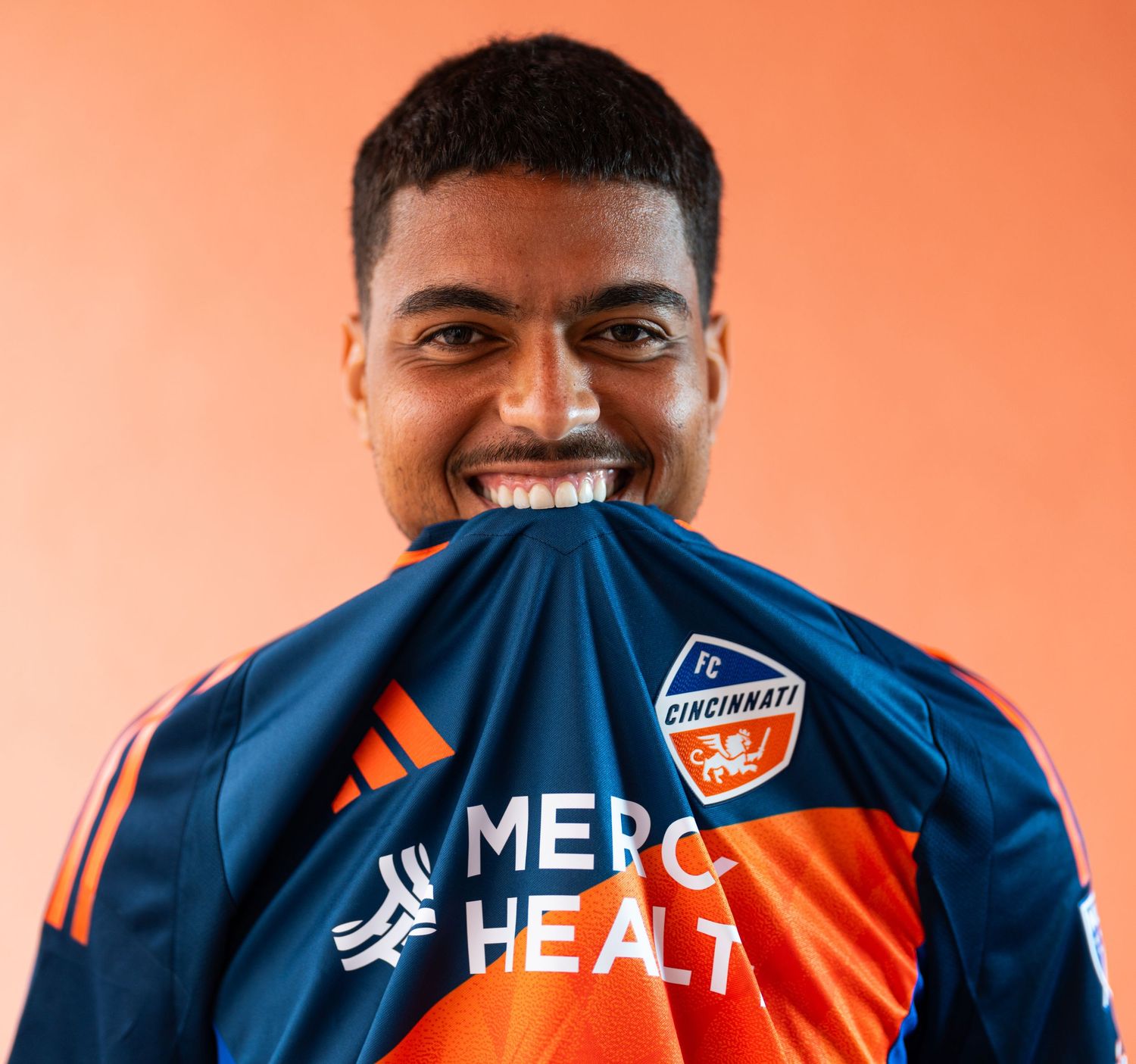 Studio portrait of a soccer player holding a jersey near their face against an orange background.
