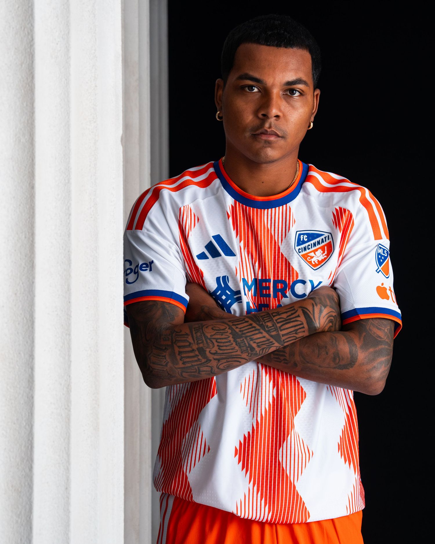 Close-up portrait of a person wearing a white and orange FC Cincinnati jersey with tattooed forearms crossed in front of a white stone colum