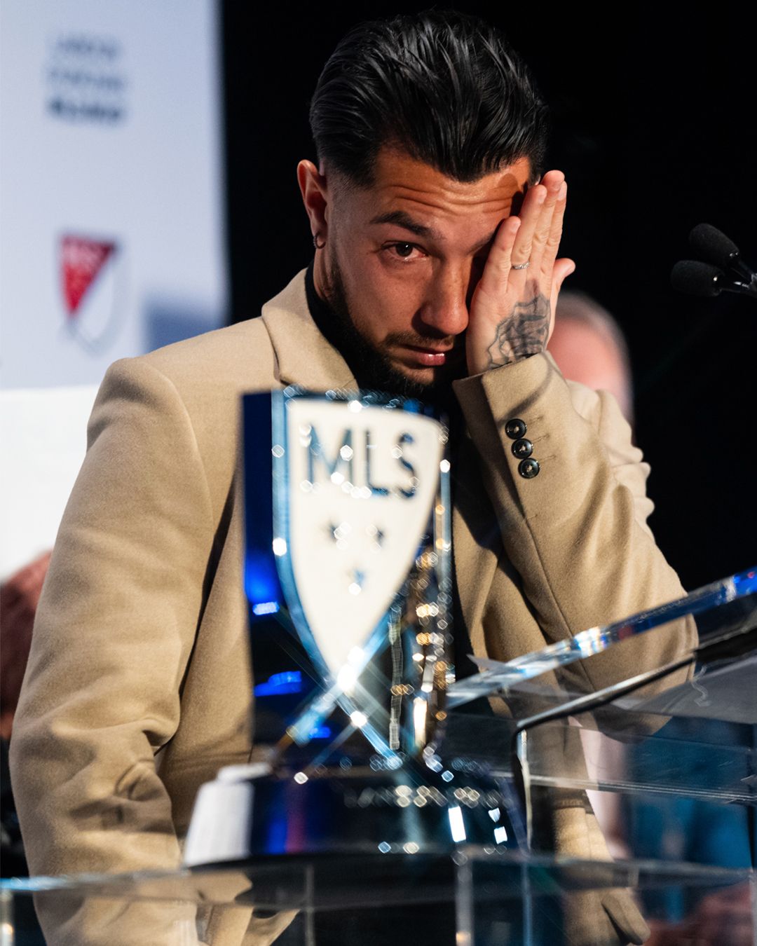 Person at a podium wipes an eye while standing beside an MLS trophy during an awards event.