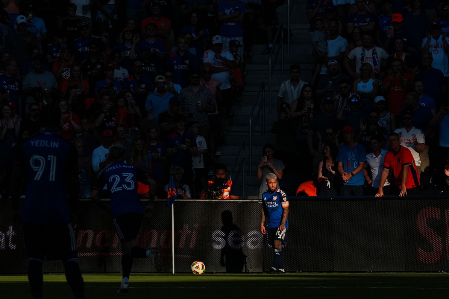 Soccer player stands near a corner kick spot illuminated by a beam of sunlight in a crowded stadium.