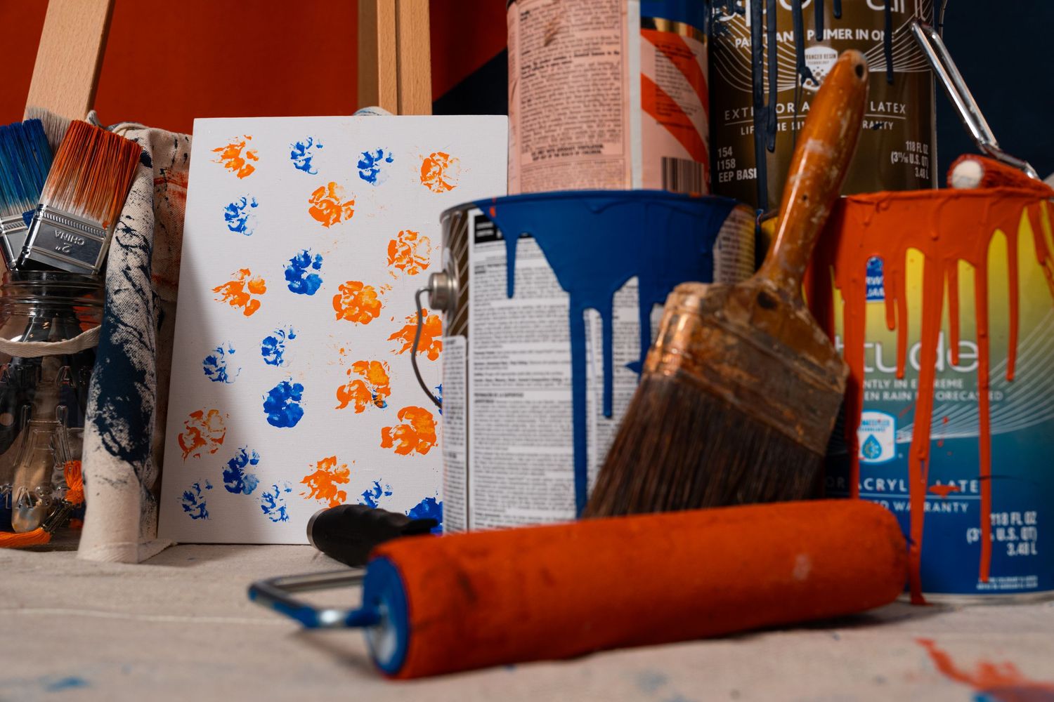 Assorted painting supplies including paint cans, brushes, rollers, and a small canvas with orange and blue paint marks.