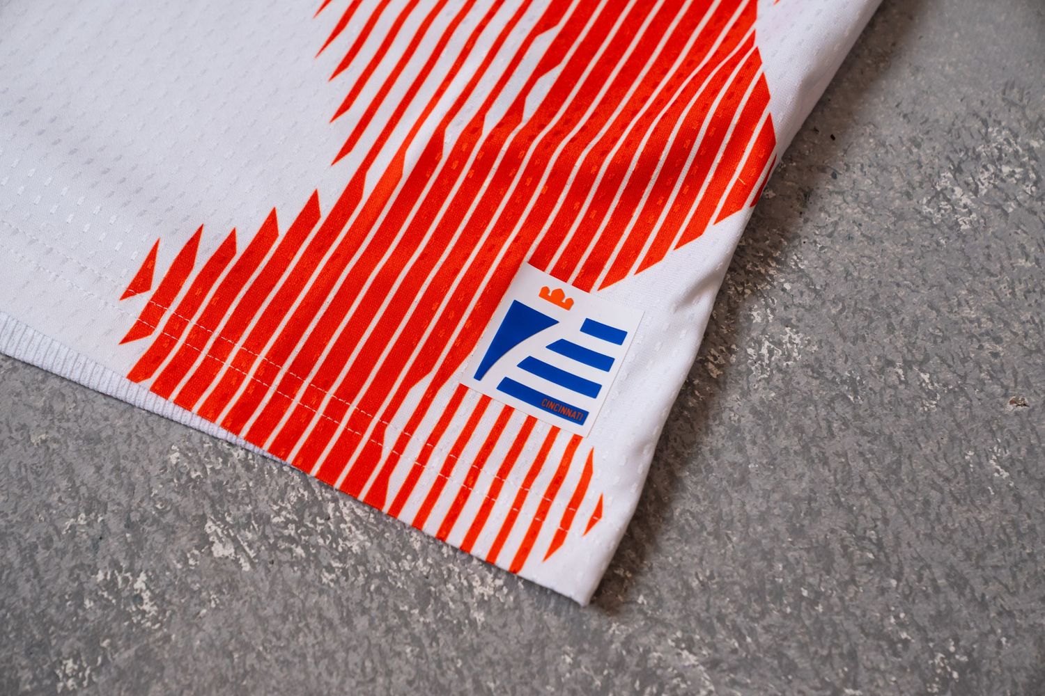 Close-up detail of the lower hem of a white and orange FC Cincinnati jersey featuring a small blue-and-orange graphic mark.