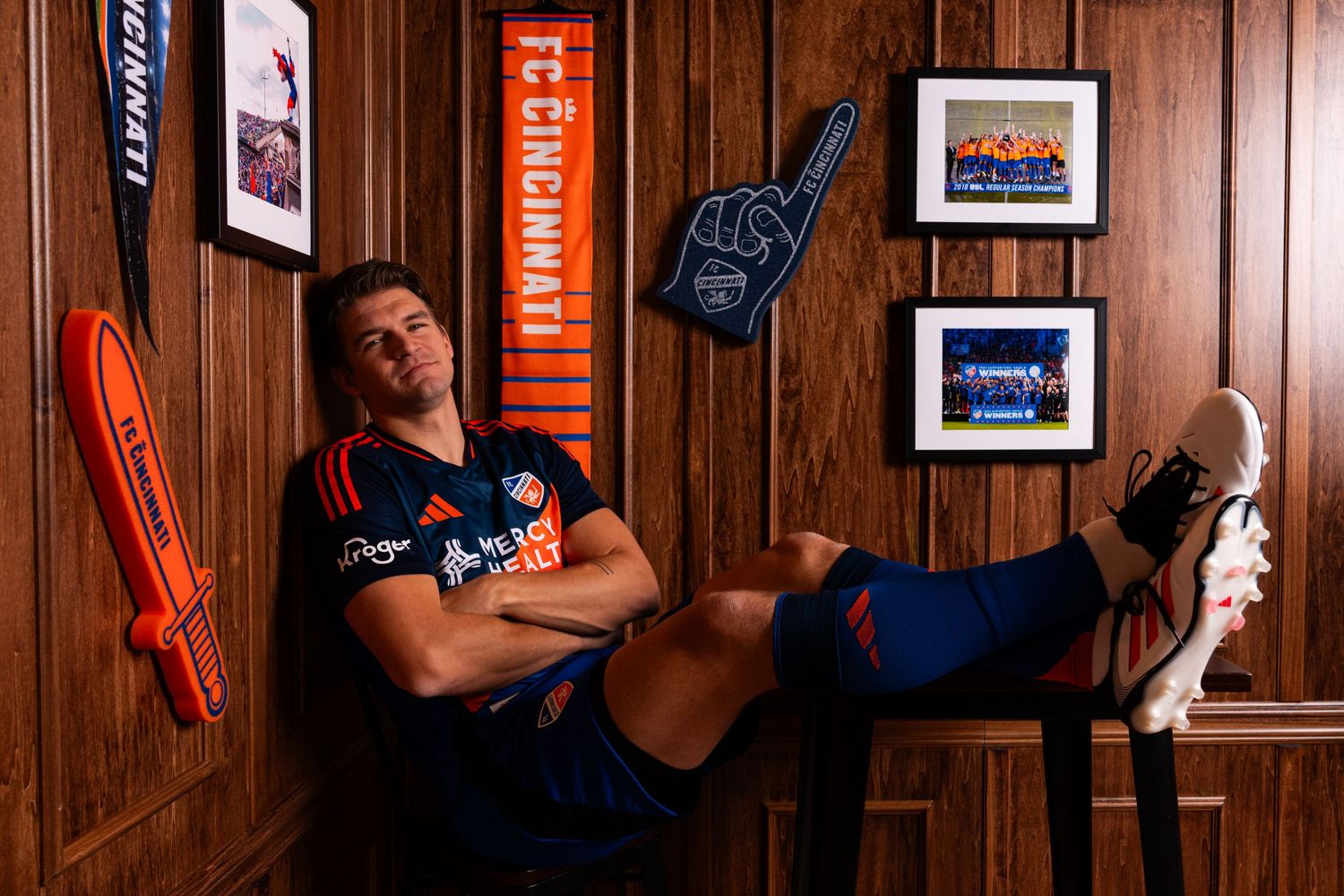 Person in a blue and orange FC Cincinnati uniform reclining in a chair with feet resting on a table inside an FC Cincinnati decorated room.