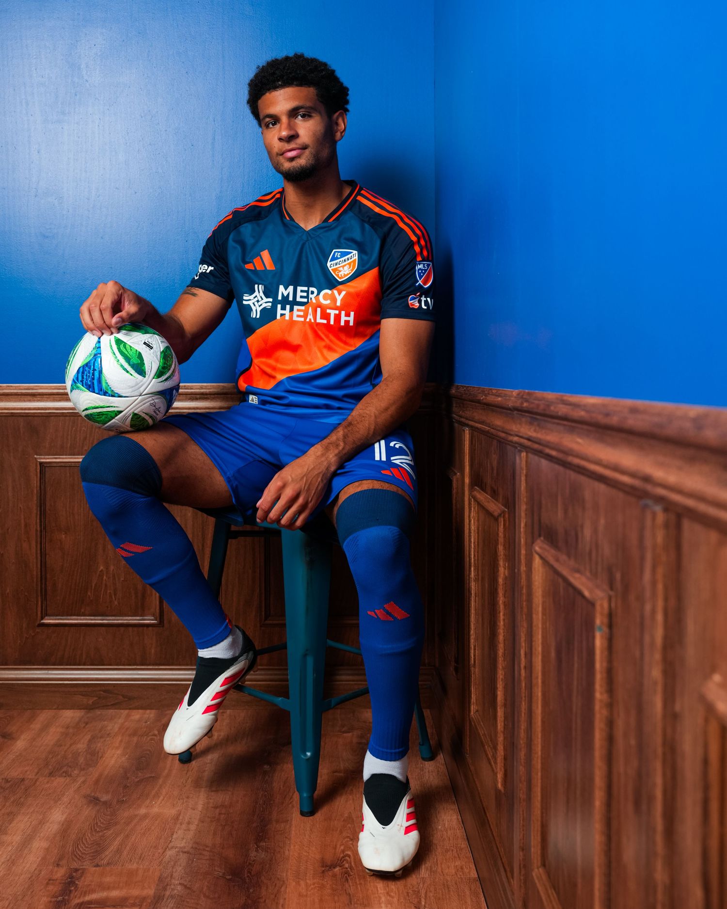 Person seated on a stool holding a soccer ball while wearing a blue and orange FC Cincinnati uniform against a blue wall.