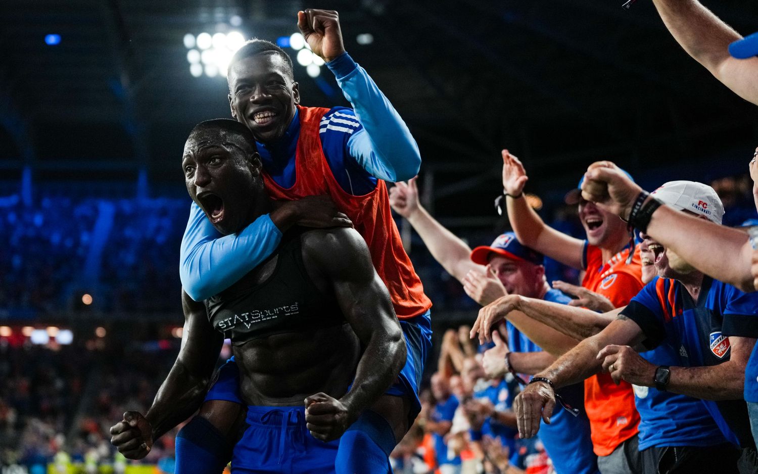 Two soccer players celebrate near supporters along the sideline during a nighttime match.