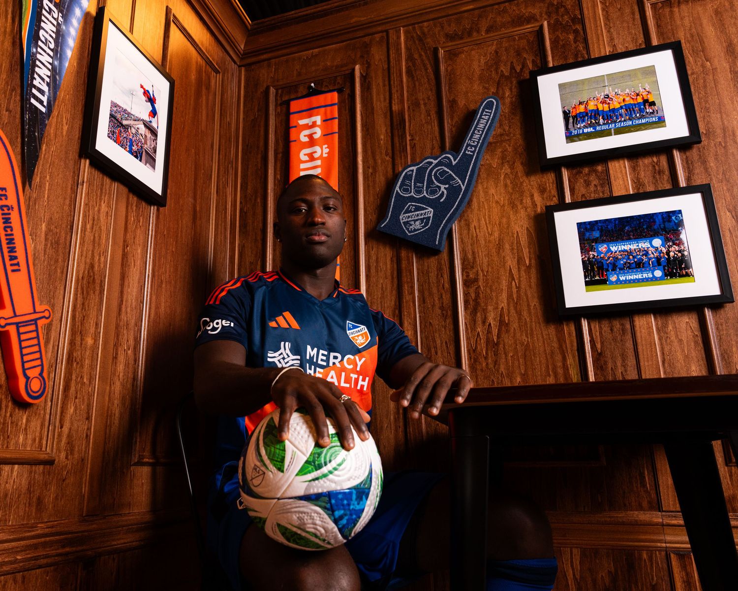 Person seated beside a table holding a soccer ball while wearing a blue and orange FC Cincinnati uniform in a wood-paneled room.