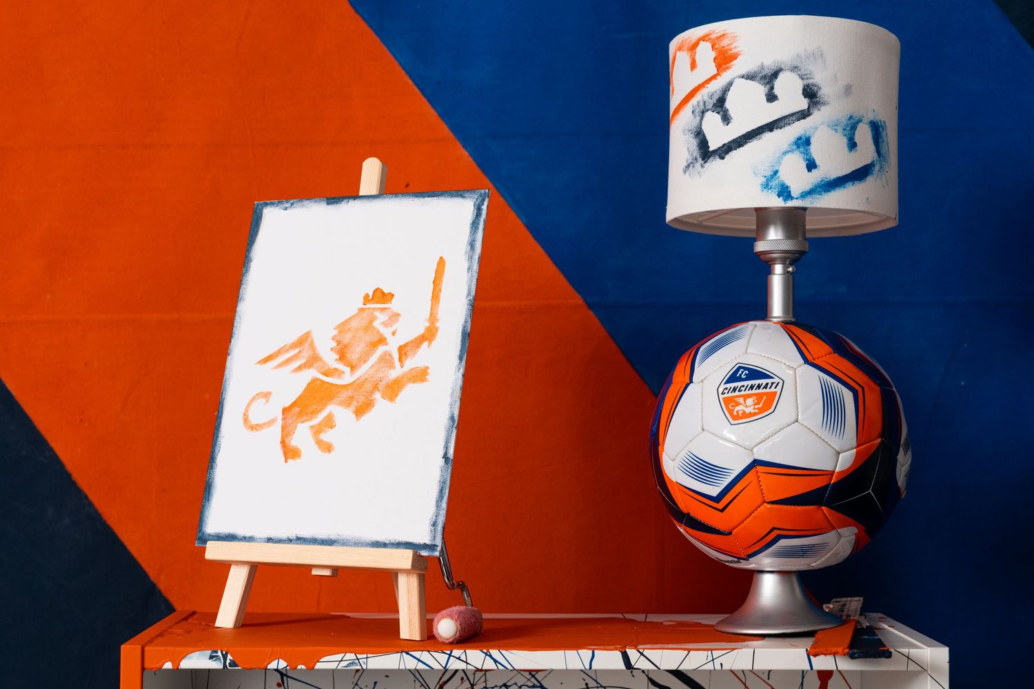 Small easel displaying a painted lion graphic beside an FC Cincinnati soccer ball mounted on a stand with a painted lampshade above it.