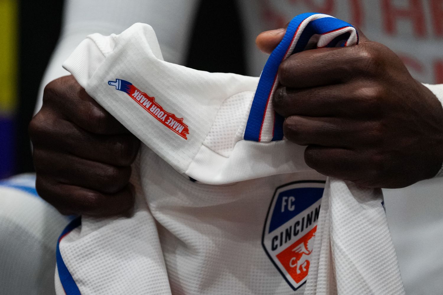 Close-up of hands holding a white FC Cincinnati jersey featuring team branding and trim details.