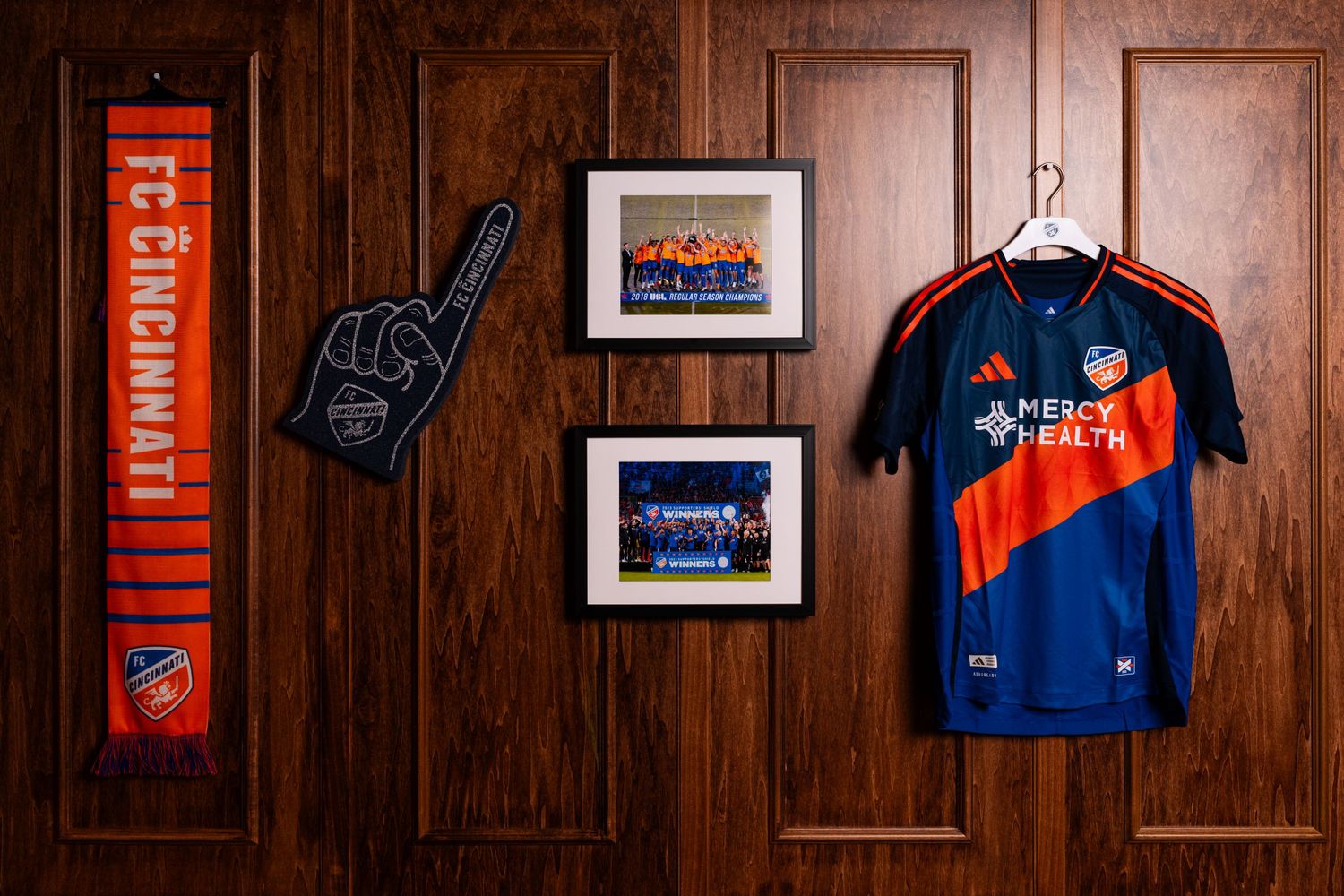 FC Cincinnati scarf, foam finger, framed photographs, and a blue and orange FC Cincinnati jersey displayed against a wood-paneled wall.