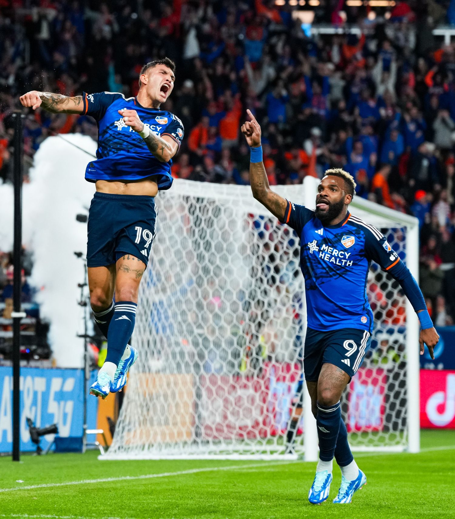 Two FC Cincinnati players celebrate in front of the goal with supporters visible in the background.