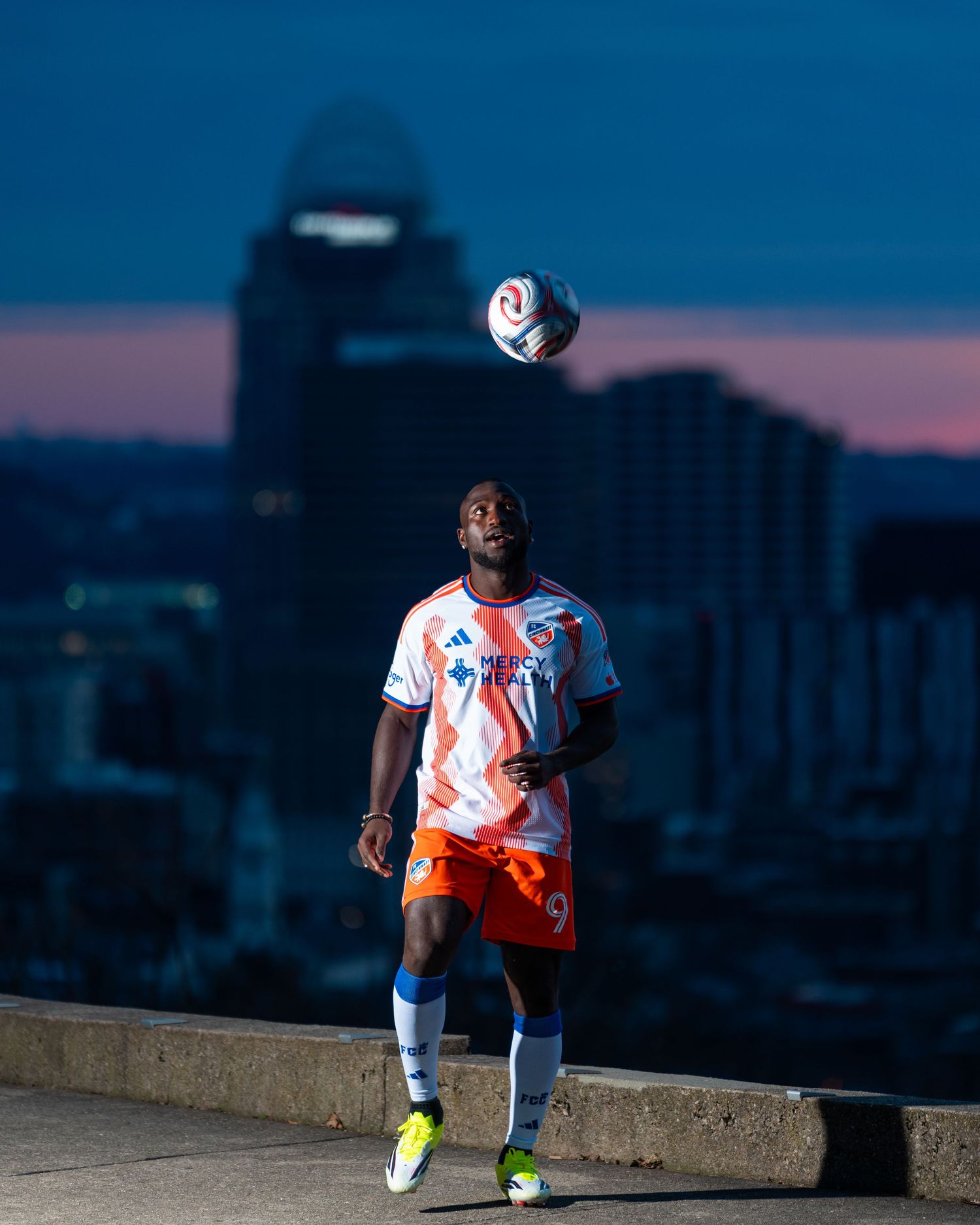 Person wearing a white and orange FC Cincinnati uniform looking upward at a soccer ball suspended above them with the Cincinnati skyline.