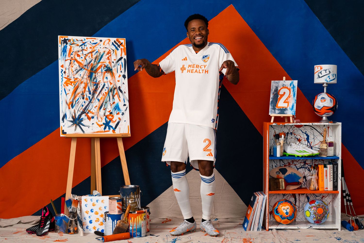 Soccer player in a white FC Cincinnati uniform posing with raised hands in an art-themed studio.