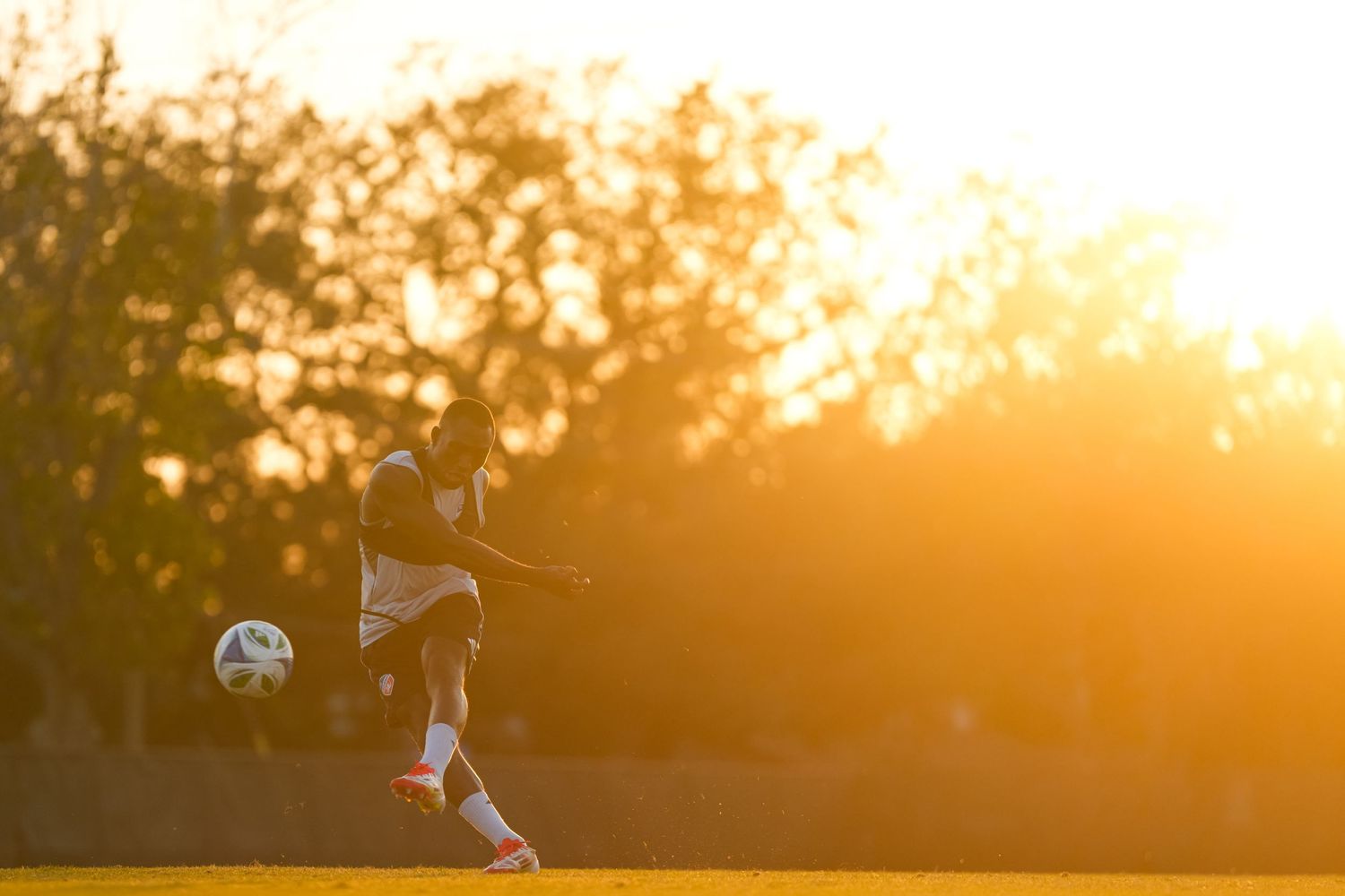Soccer player kicks a ball during training at sunset with warm golden light across the field.
