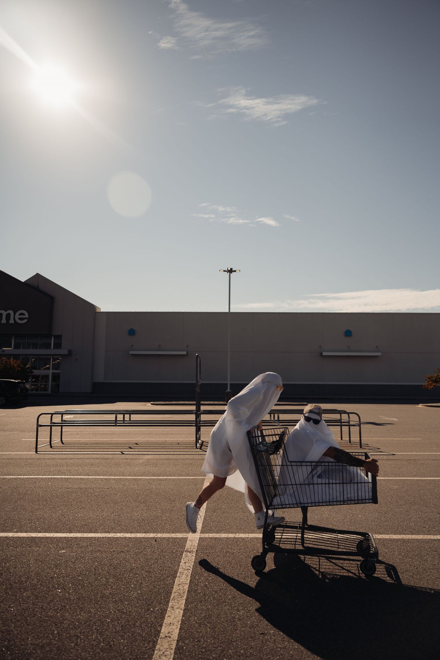 Person in ghost costume pushes another ghost in a shopping cart across a sunny parking lot.