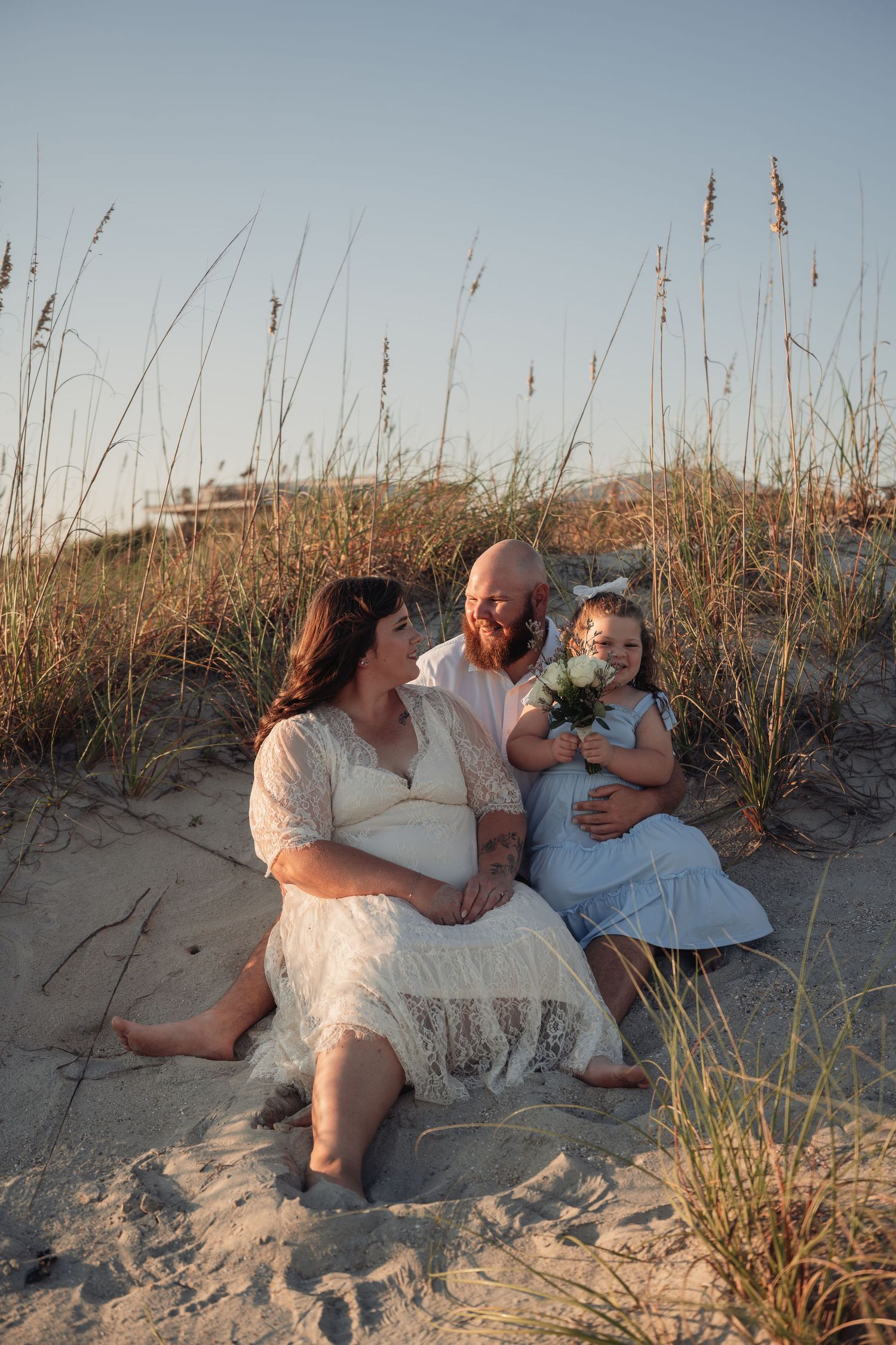 Family of three sitting together on a sandy beach dune with sea oats at sunset, a young girl holding flowers between her smiling parents.