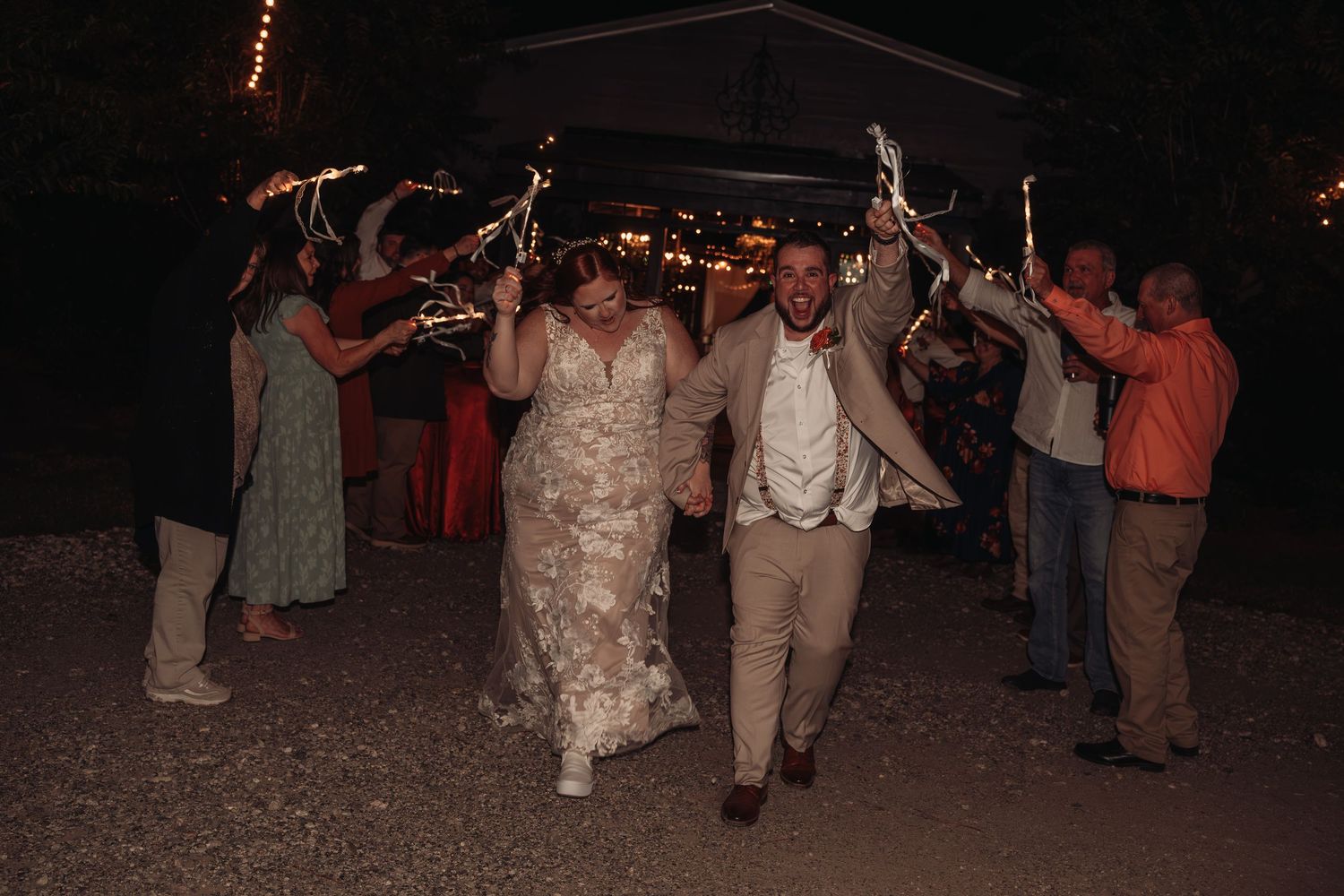 Newlyweds walk hand in hand through a nighttime sparkler send-off as wedding guests cheer along the path.