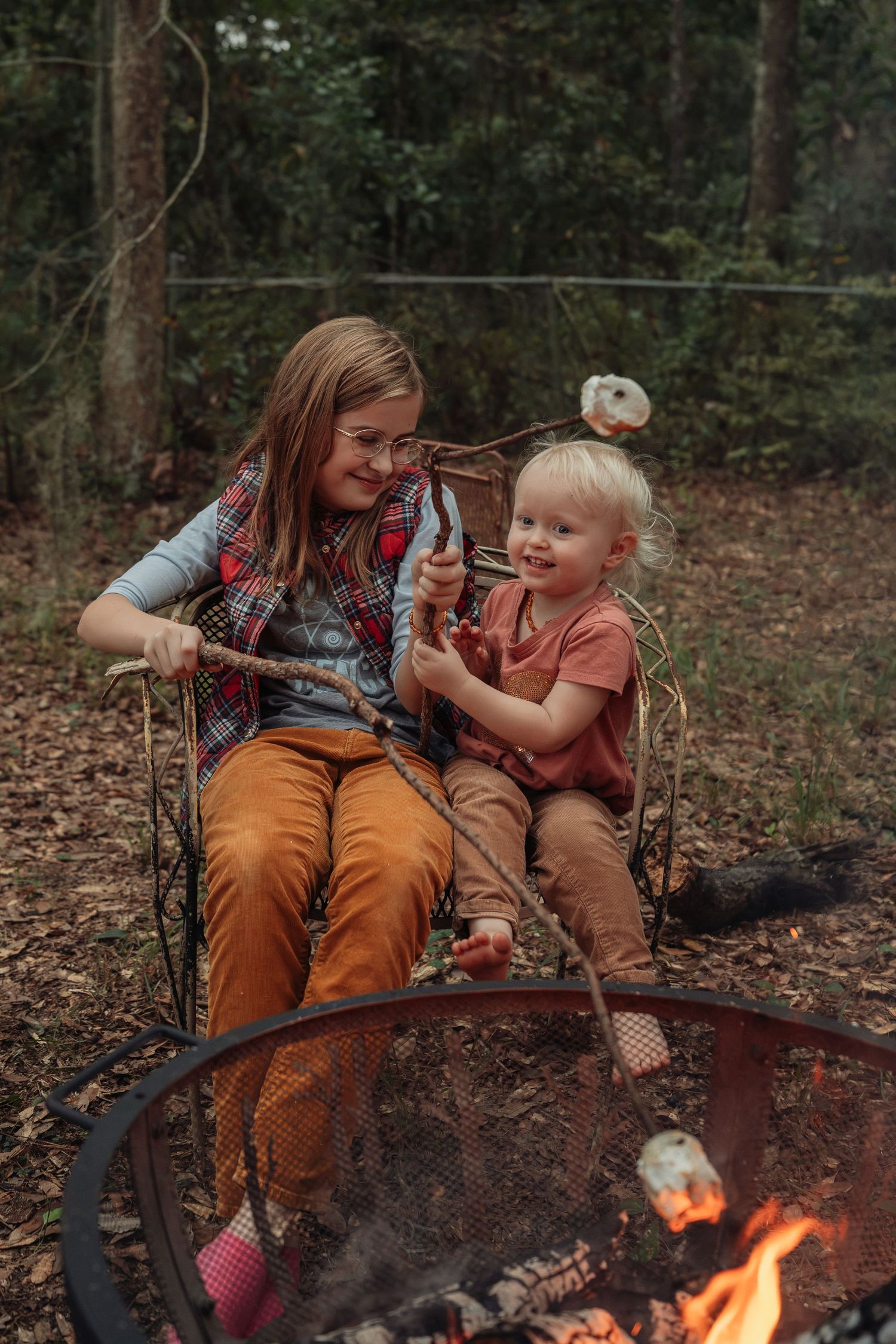 Two kids sitting together by a campfire in the forest, roasting marshmallows on sticks while smiling.
