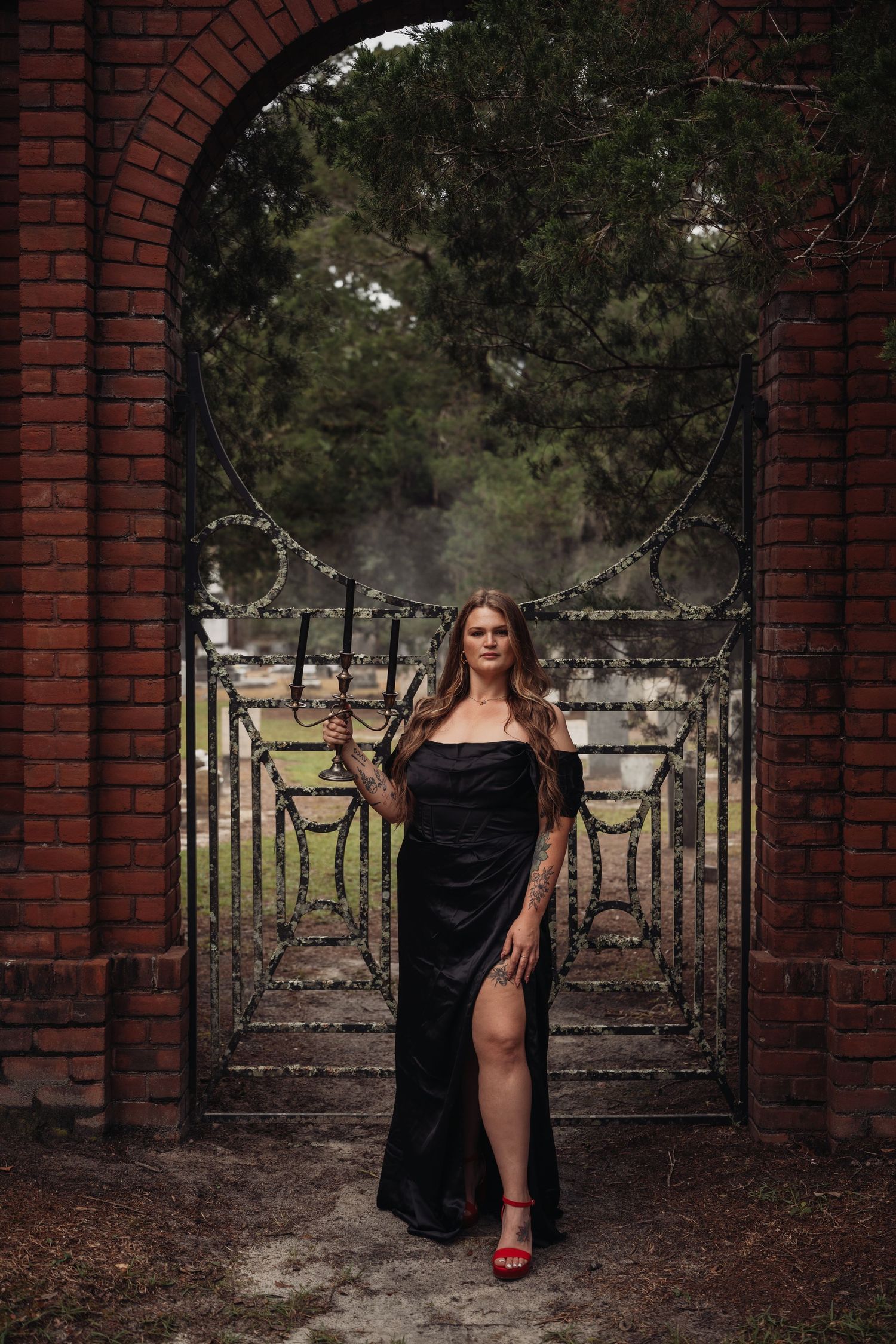 Woman in black off-shoulder gown with red heels poses before ornate iron gates framed by brick archway.