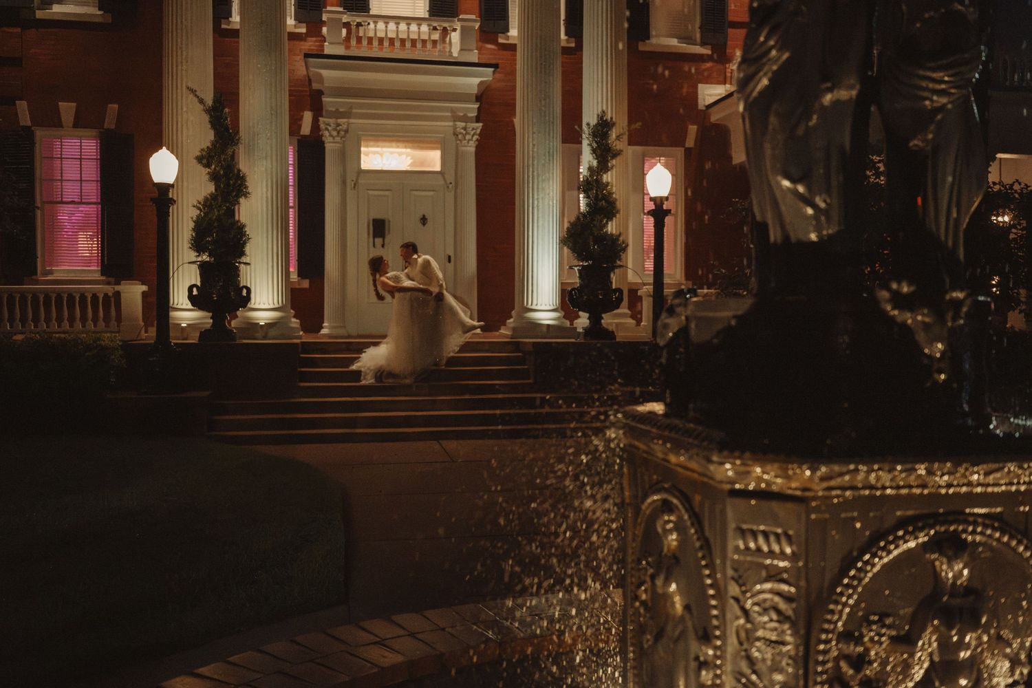 Elegant wedding couple dancing on grand mansion steps at night with ornate golden fountain in foreground.