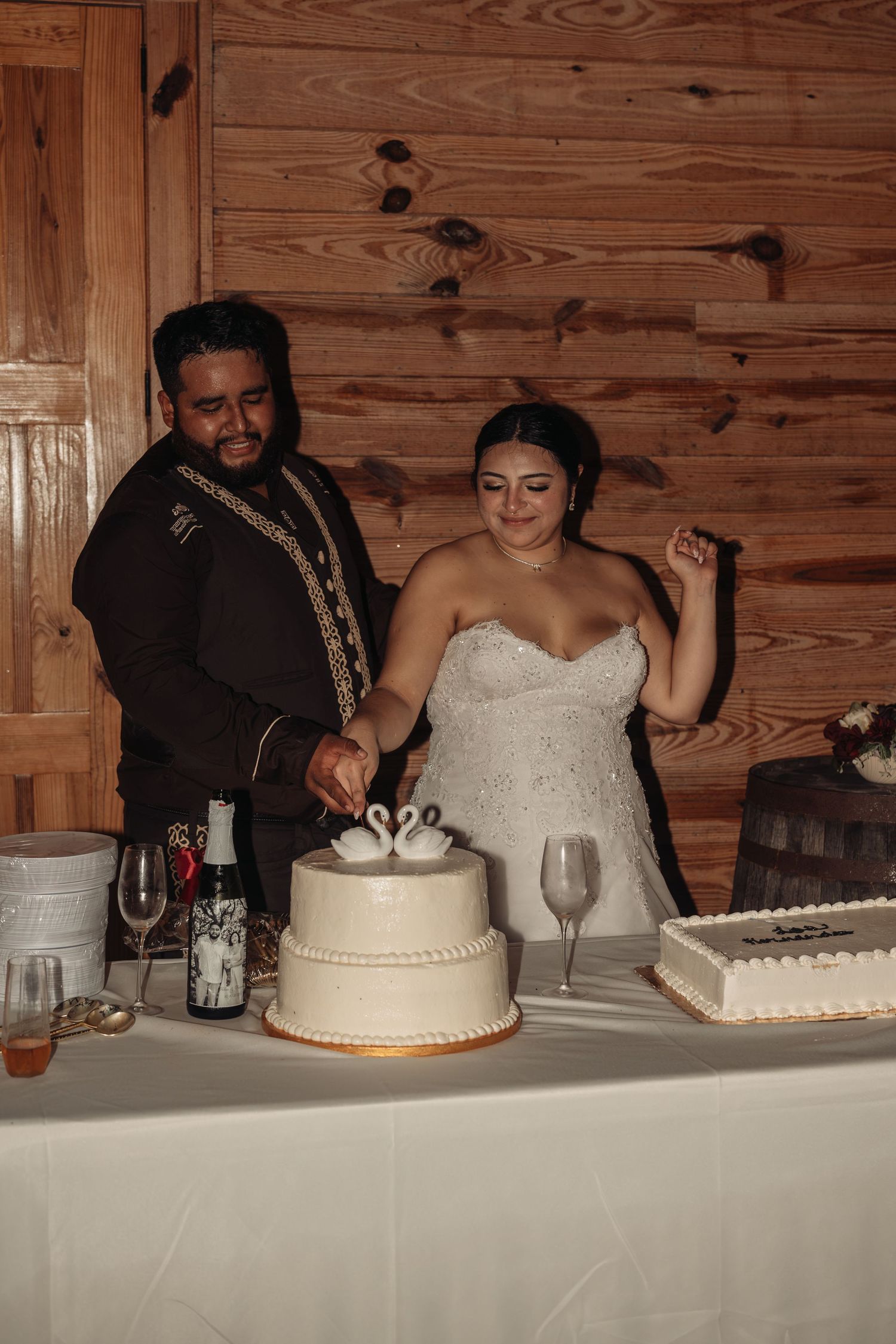 Bride and groom cutting their two-tier wedding cake decorated with swan toppers at a rustic wood-paneled venue.