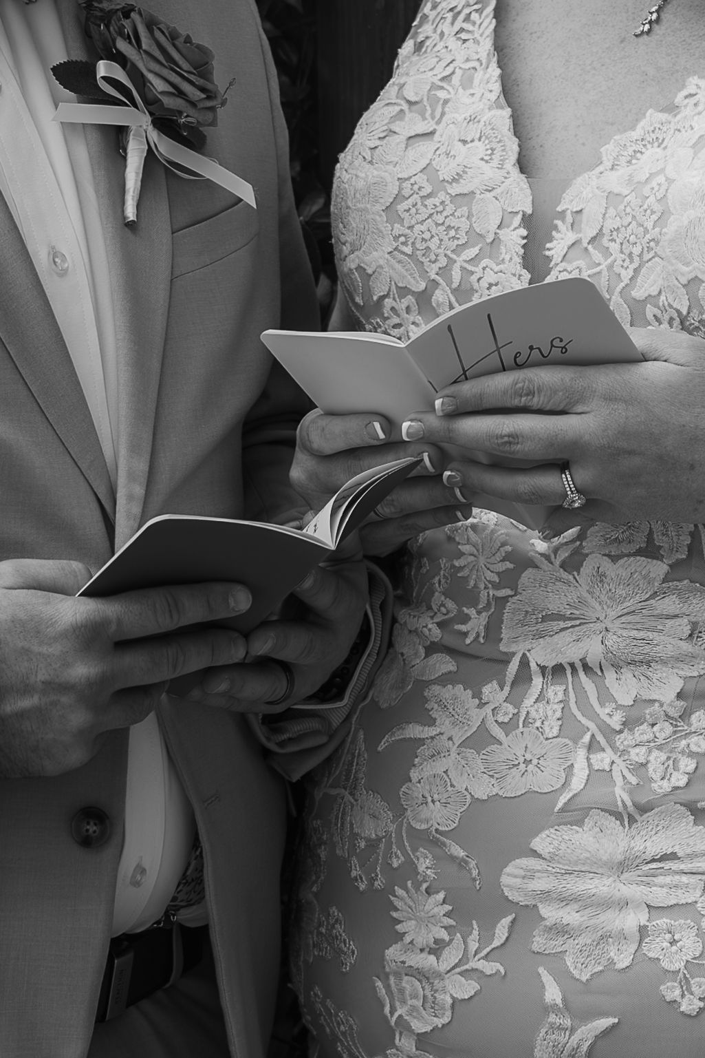 Bride in a lace wedding dress and groom in a suit with a boutonniere read their handwritten vows from small booklets in black and white.