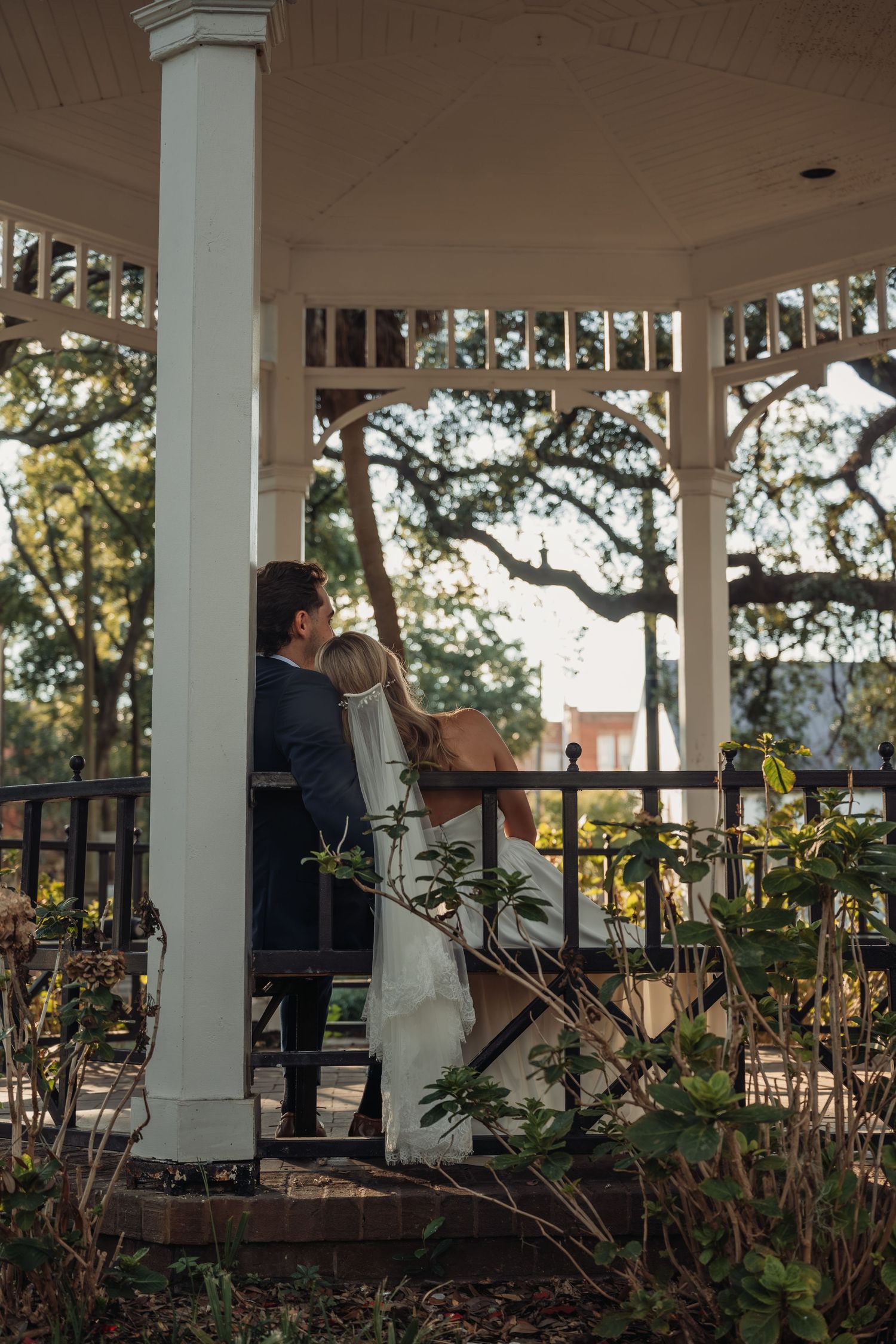 Bride and groom sit together in a white gazebo surrounded by lush greenery and oak trees.