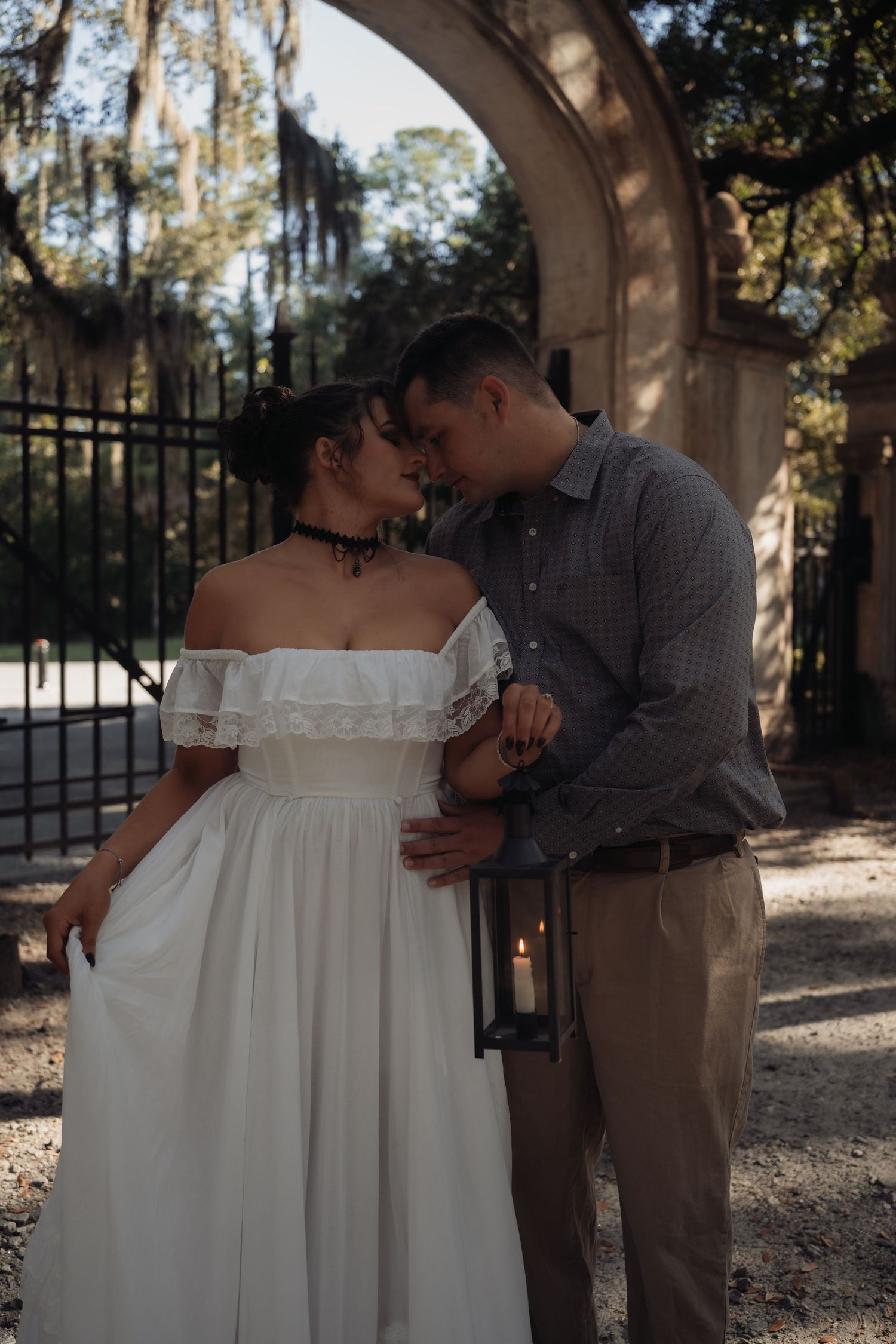 Romantic couple sharing an intimate moment by an iron gate, woman in white off-shoulder gown, man holding a glowing lantern.