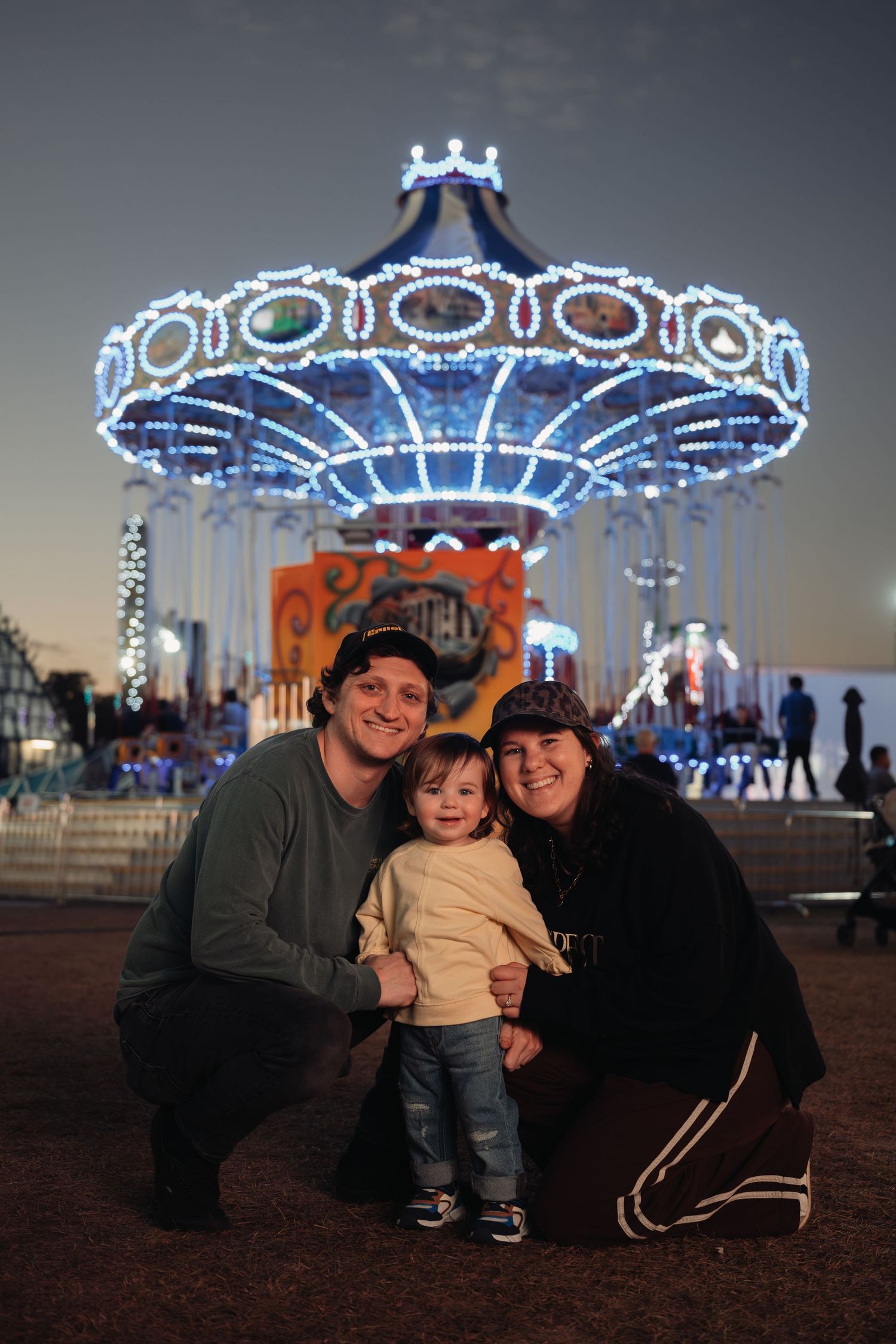Smiling couple kneeling with their toddler in front of a glowing carousel lit with blue lights at an evening fair.