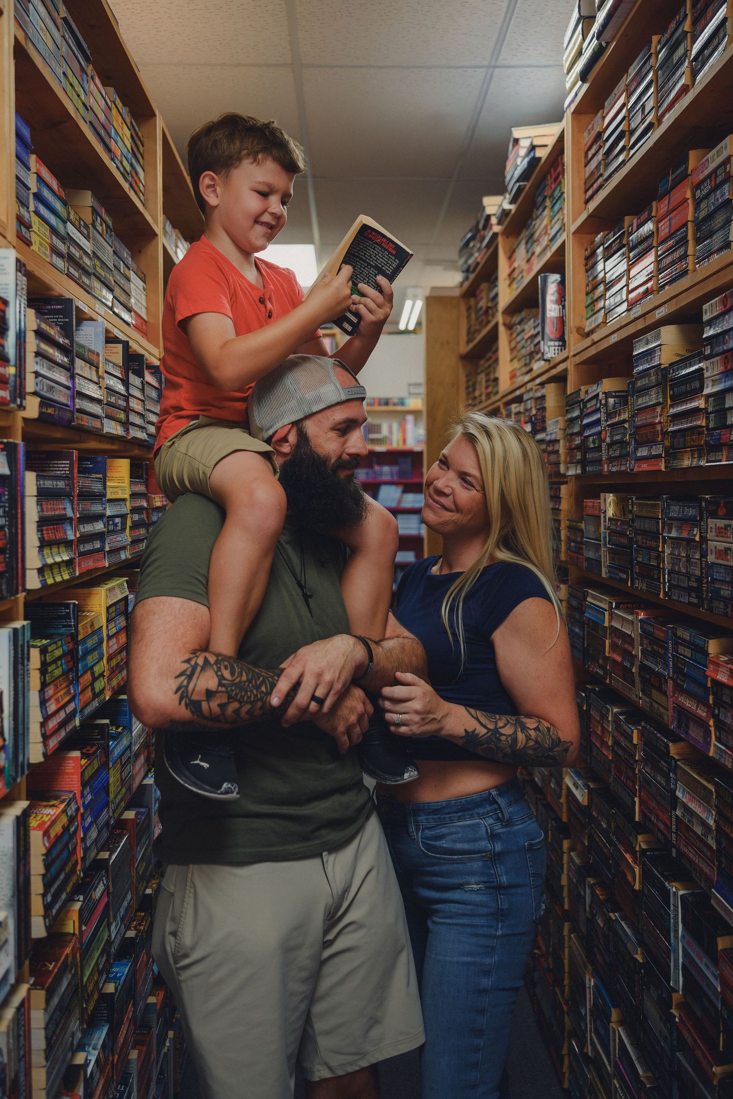 Family with tattooed parents and young boy on dad's shoulders browsing books in a library aisle.