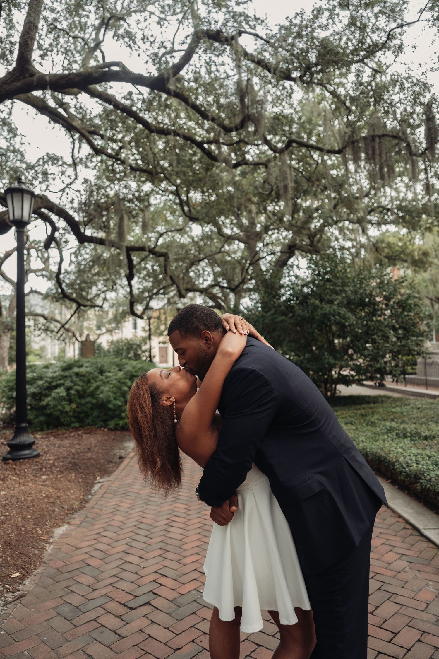 Couple sharing a passionate dip kiss on brick pathway surrounded by moss-draped oak trees in Savannah.