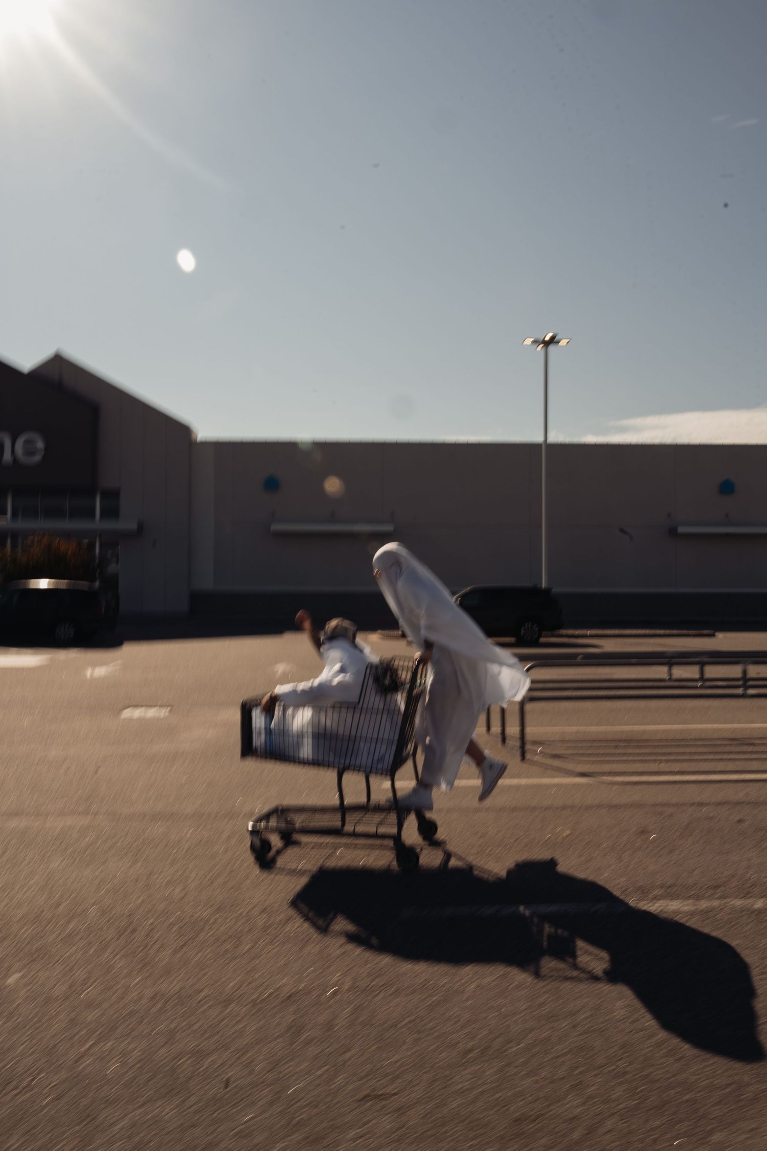 Person in white ghost costume pushes another ghost in a shopping cart across a sunlit parking lot.