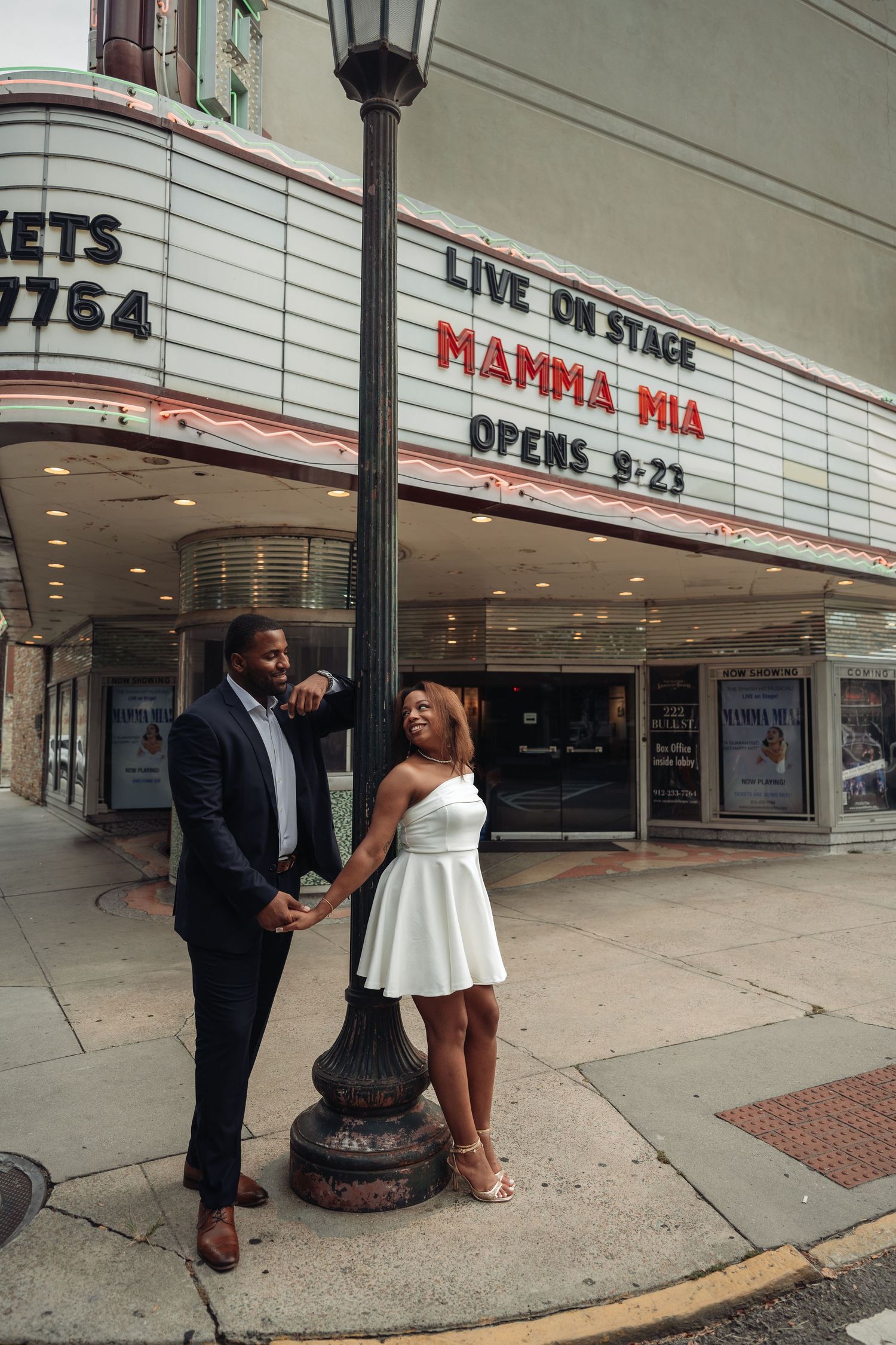 Elegant couple holding hands by a vintage street lamp outside a theater showing Mamma Mia, woman in white dress, man in suit.