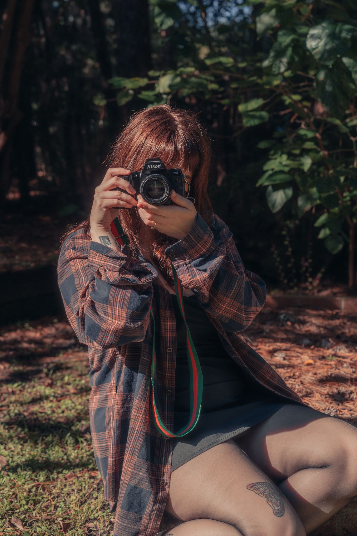 A redhead woman in a plaid shirt sits outdoors, holding a Canon camera up to her face, with a tattoo visible on her leg.