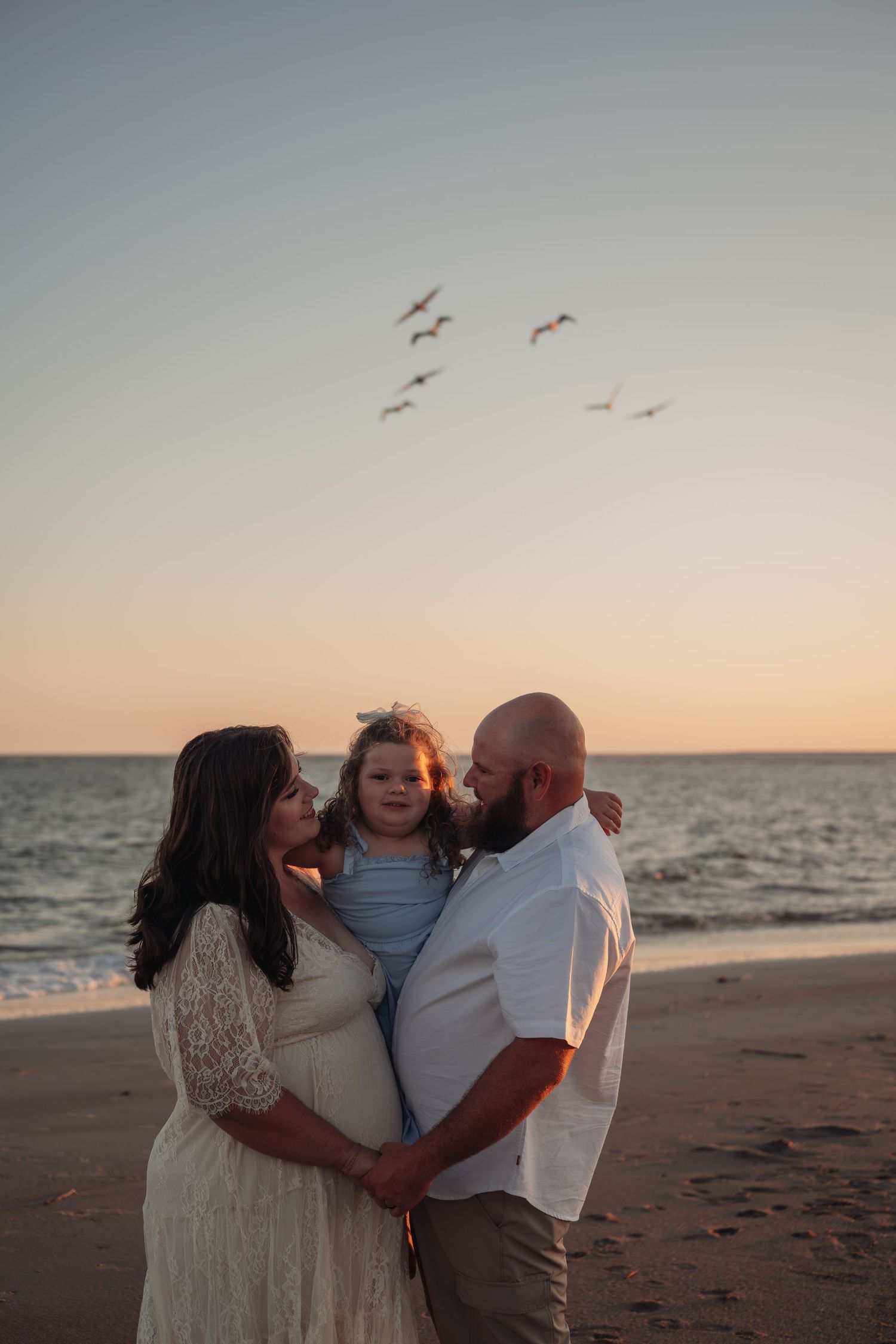 Expecting couple in a lace dress and white shirt hold their young daughter on a beach at sunset as seagulls fly overhead.