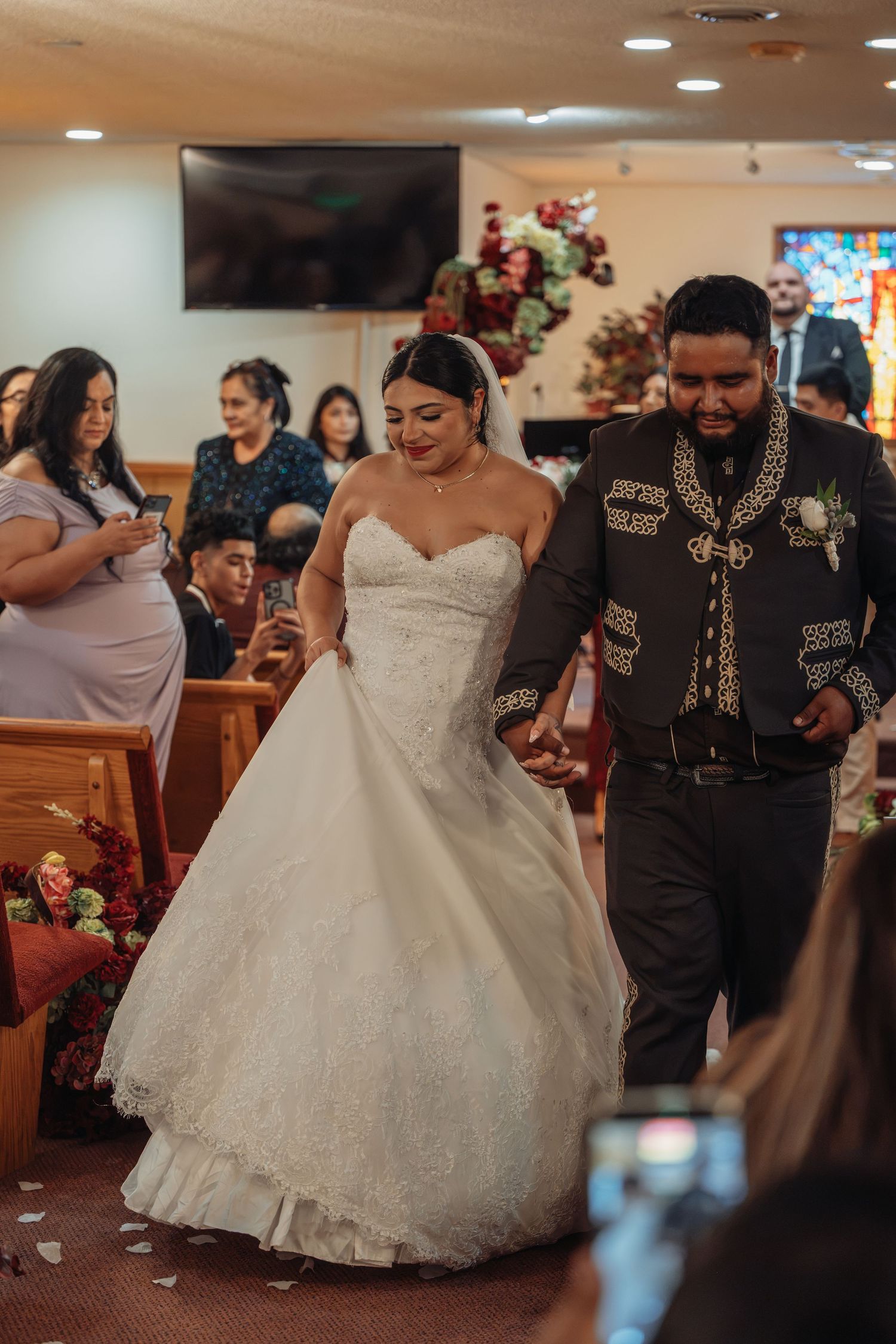Bride in white ball gown and groom in black mariachi suit walk down church aisle at wedding ceremony.