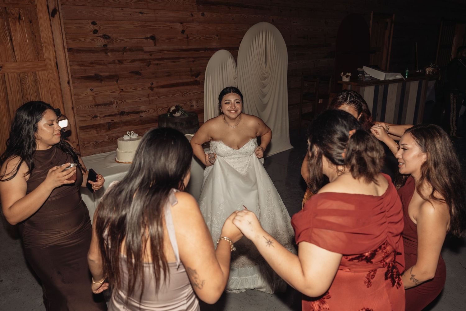 Bride in white gown surrounded by bridesmaids in a rustic barn setting during wedding celebration.