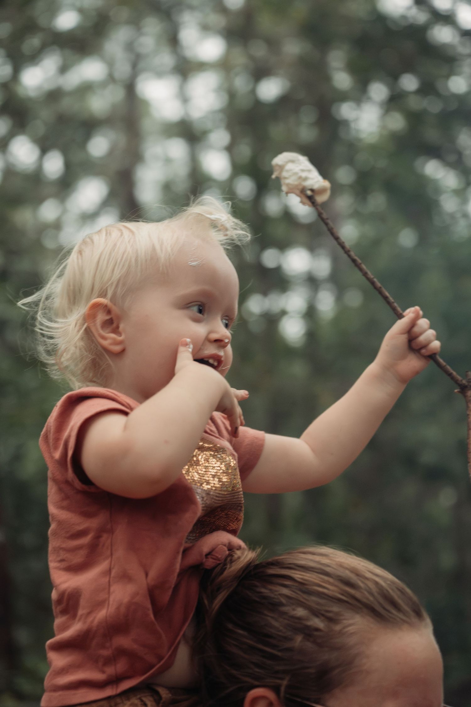 Toddler with blonde hair sits on shoulders holding a marshmallow on a stick outdoors in a forest setting.