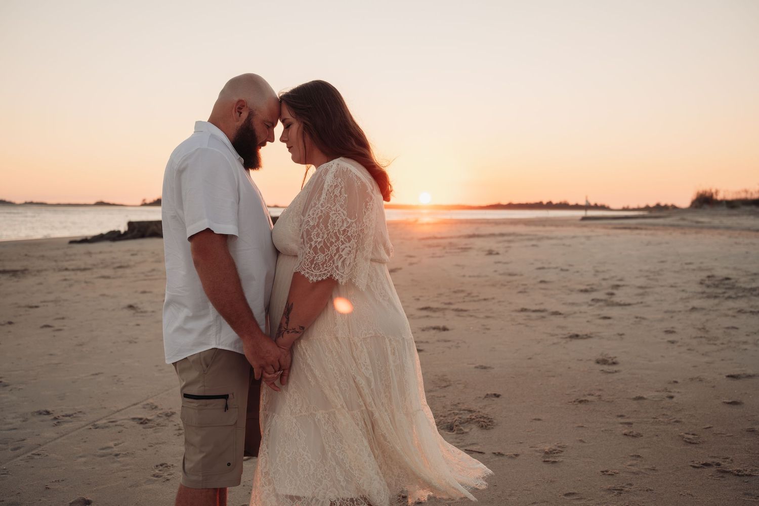 Couple standing forehead to forehead holding hands on a sandy beach at sunset, the woman in a flowing cream lace dress.