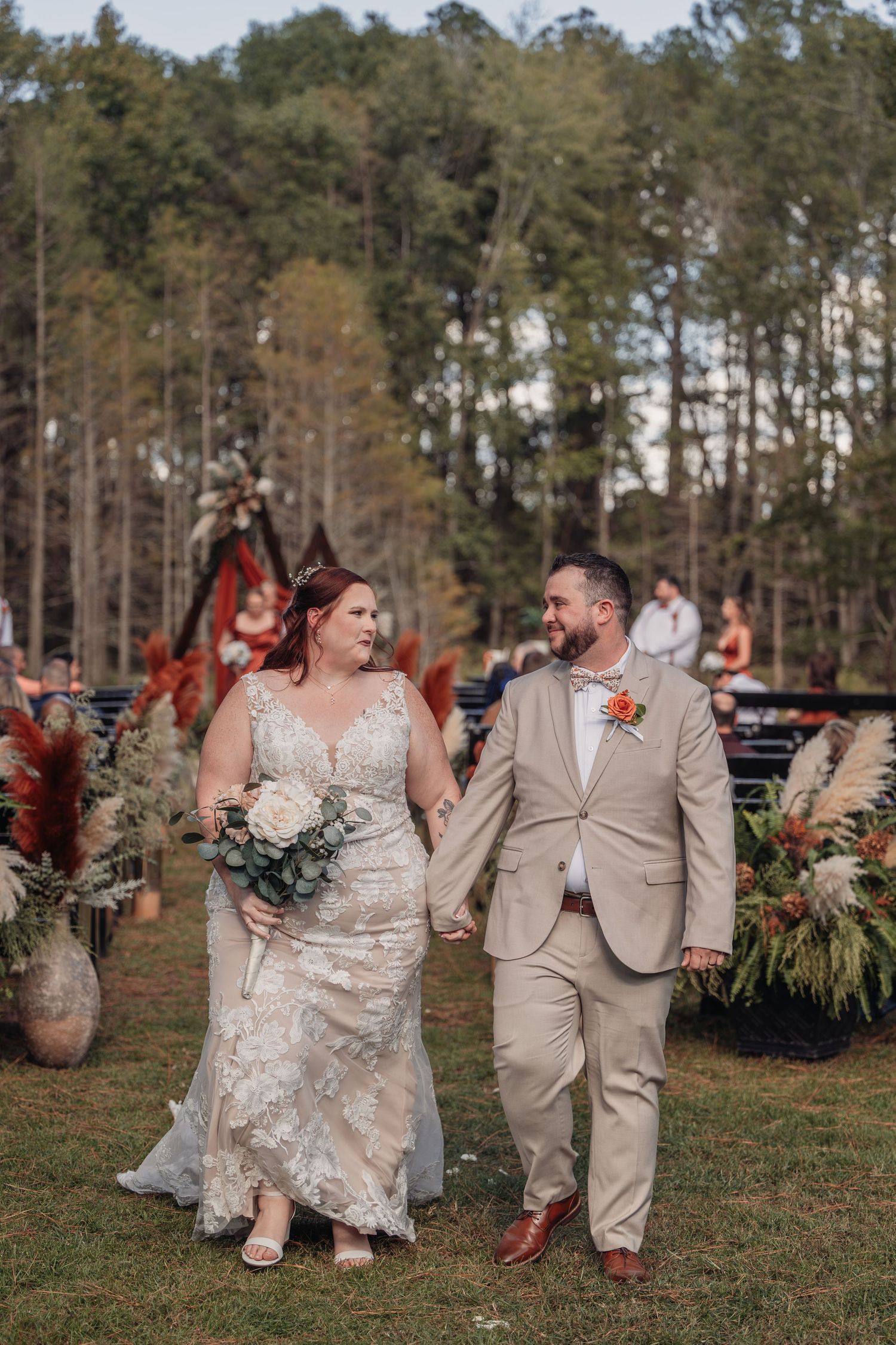 Smiling bride in a lace gown and groom in a tan suit hold hands walking down an outdoor wedding aisle lined with pampas grass.