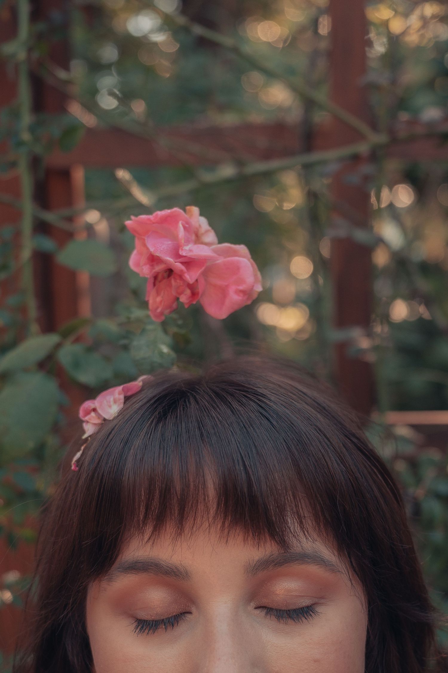 Woman with closed eyes and dark bangs wearing a pink rose in her hair, set against a lush green garden backdrop.