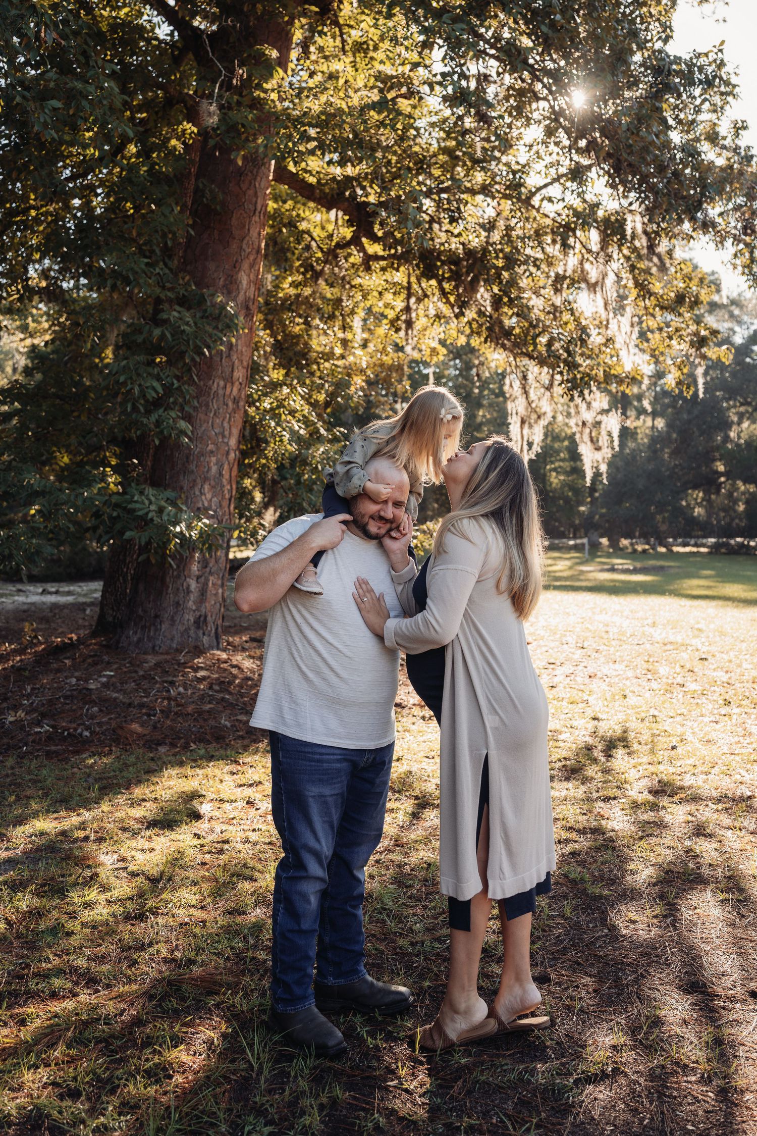 A family of three shares a warm embrace in a sunlit park, with a child sitting on dad's shoulders as mom leans in.
