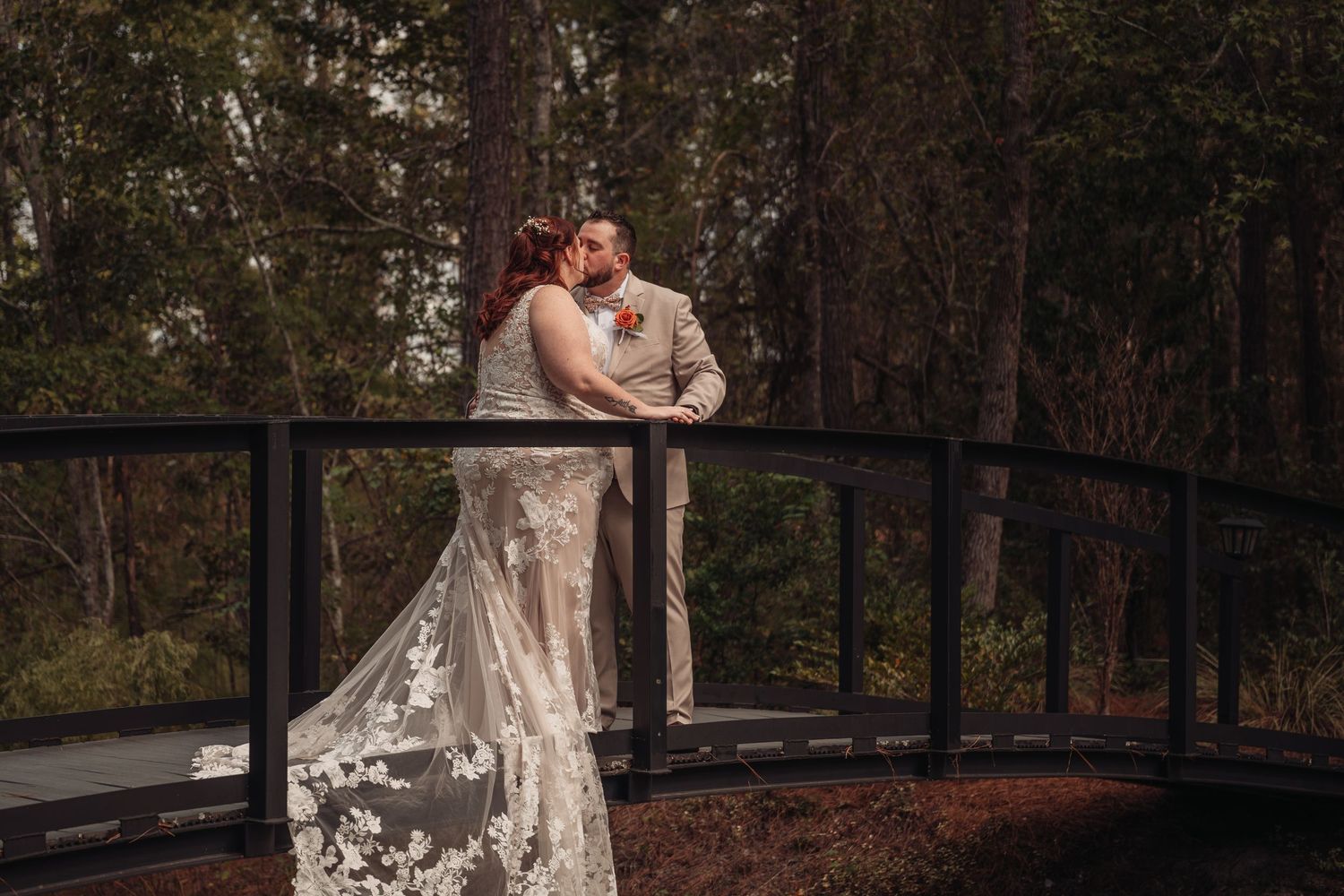 Bride in a lace wedding gown and groom in a tan suit share a kiss on a dark wooden bridge surrounded by a lush, moody forest.