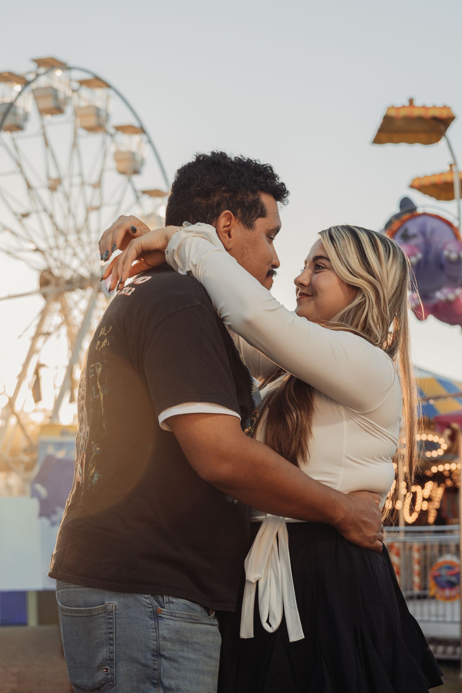 Smiling couple embracing at a carnival at sunset, with a glowing Ferris wheel and colorful fair rides in the background.