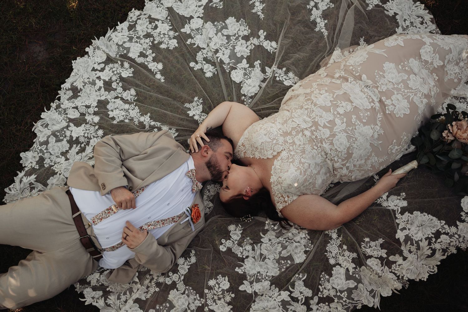 Overhead view of a bride and groom lying on the grass, sharing a tender kiss on a wide, lace-appliqué wedding gown train.