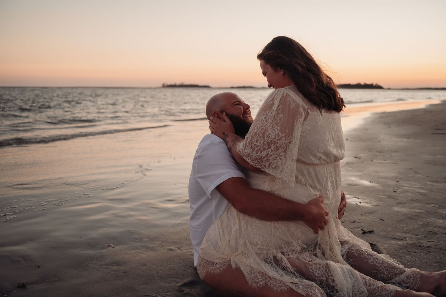 Couple embracing on a sandy beach at sunset, the woman in a flowing cream lace dress sitting close to her partner by the calm ocean.