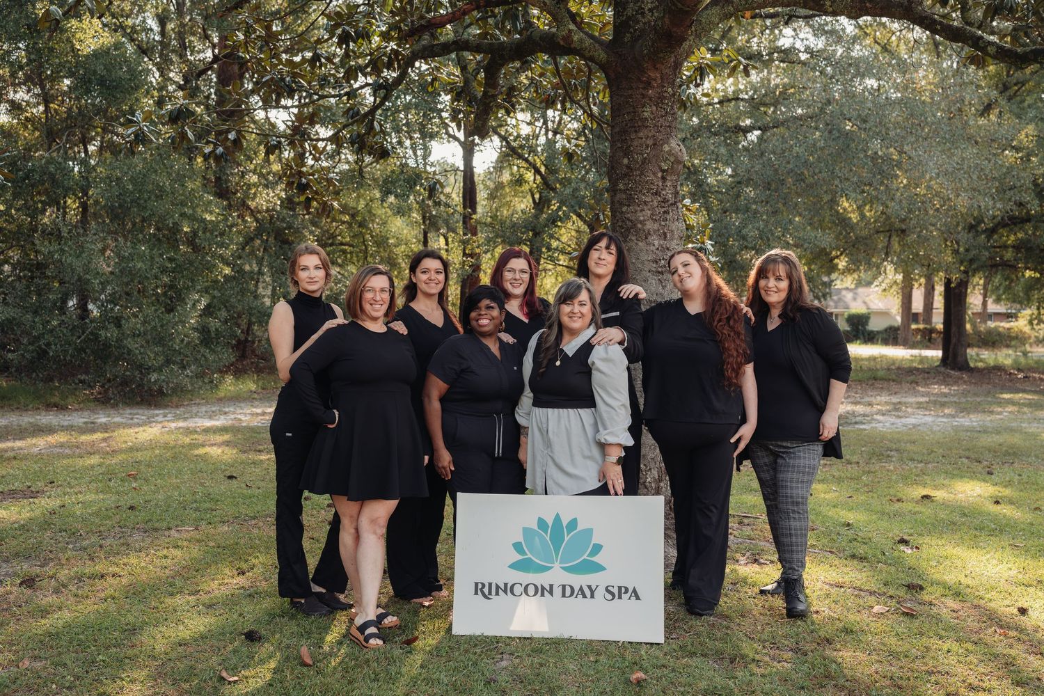 Group of nine smiling women in black attire stand outdoors under oak trees behind a Rincon Day Spa sign with a teal lotus logo.